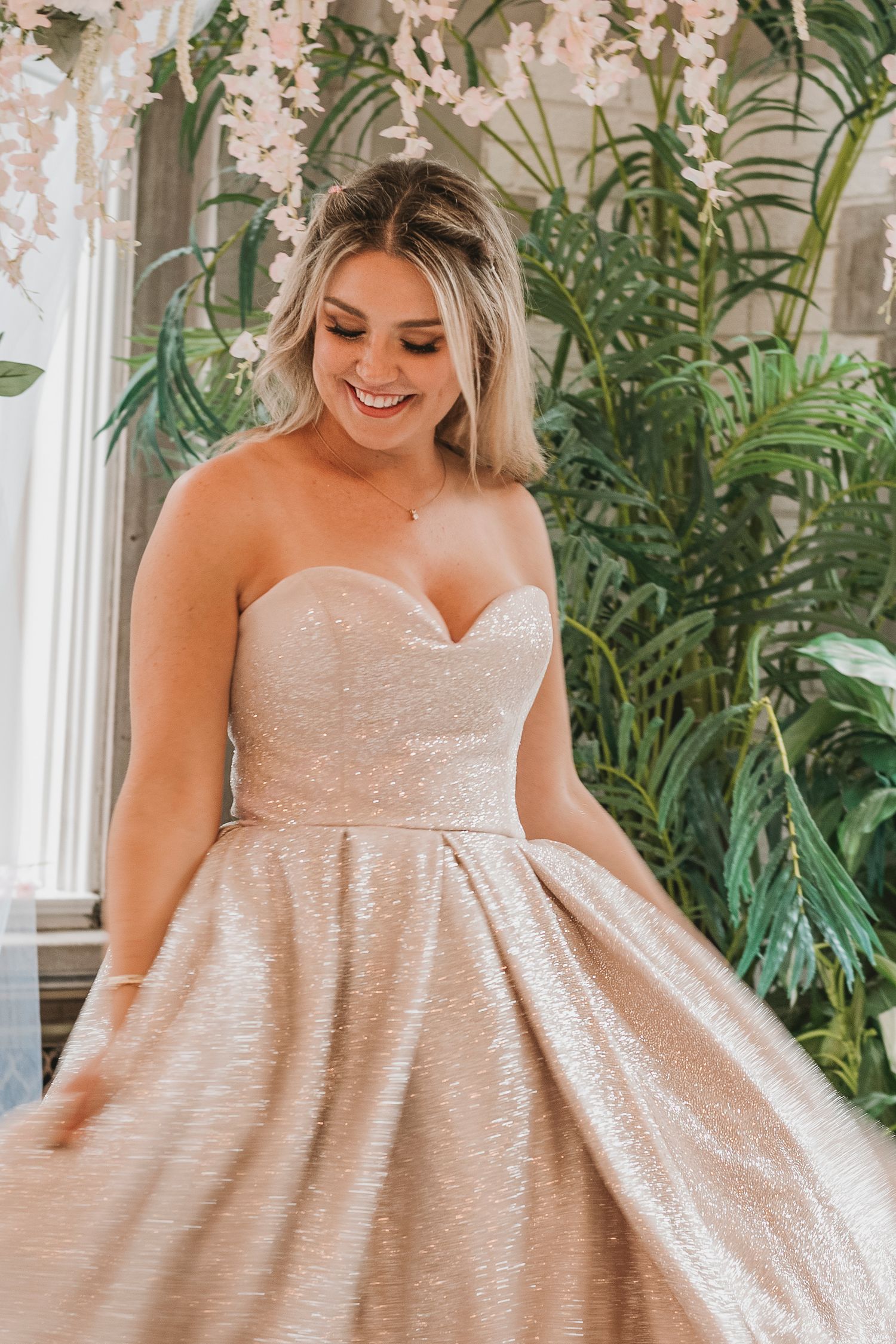 Smiling woman in strapless rose gold ballgown surrounded by tropical greenery and white flowers.