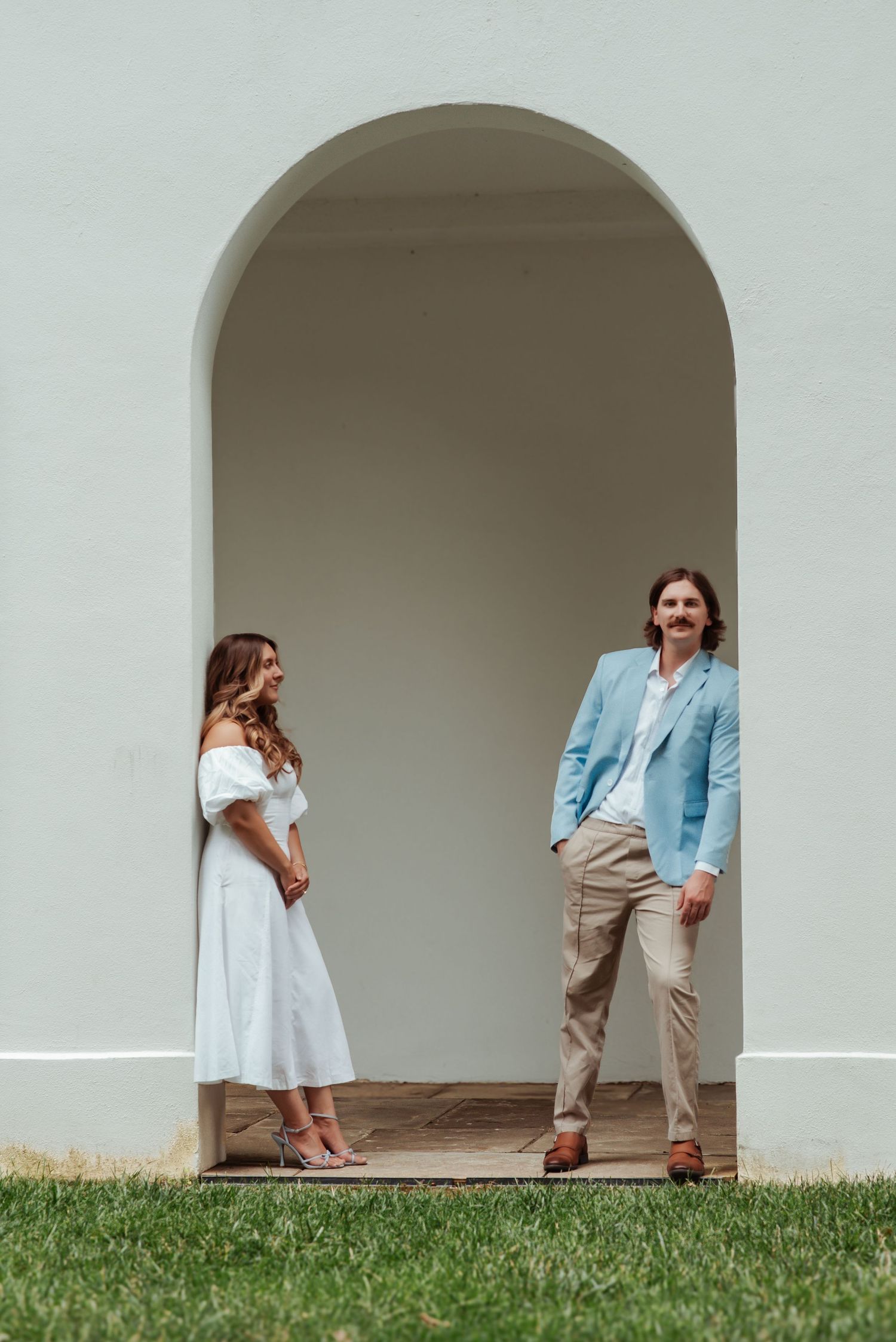 Couple standing apart under a white arched doorway, woman in white dress, man in light blue blazer.