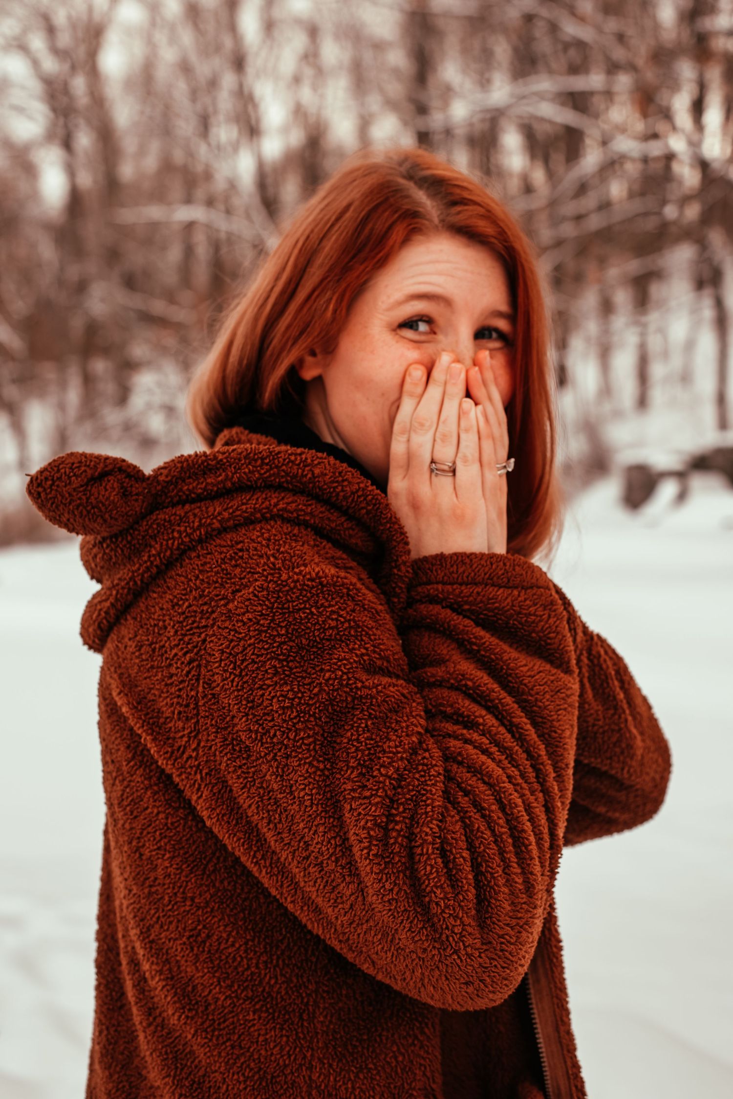 A redhead woman in a brown teddy coat covers her face with her hands while laughing in snow.