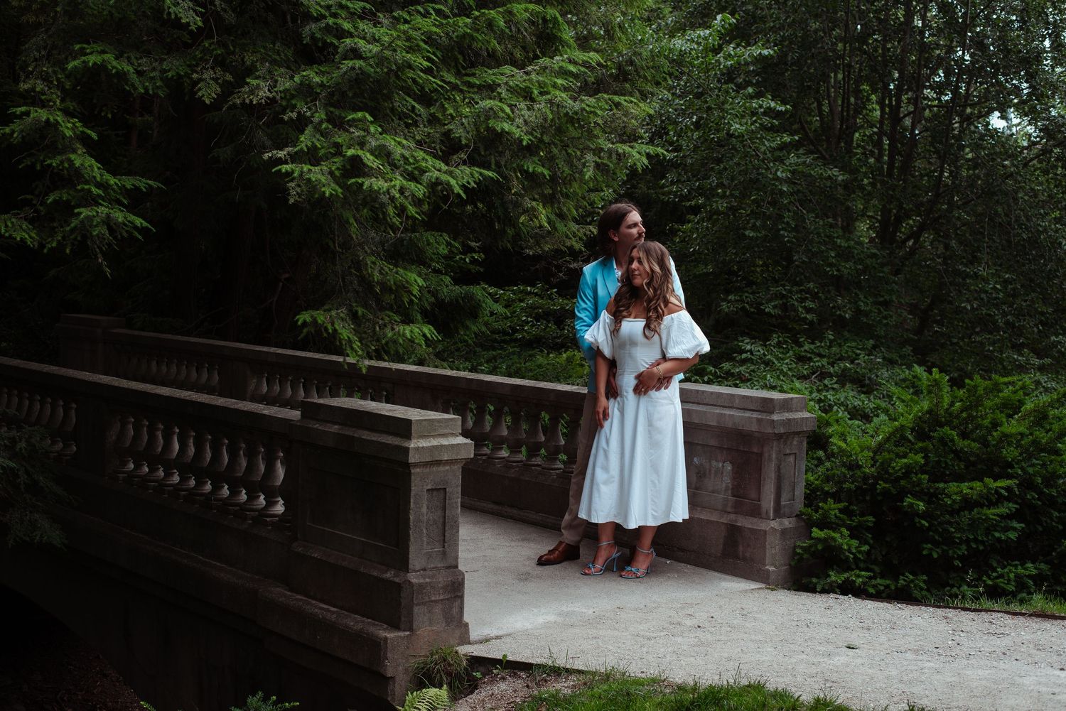 Couple embracing on a stone bridge surrounded by lush green trees in a park setting.
