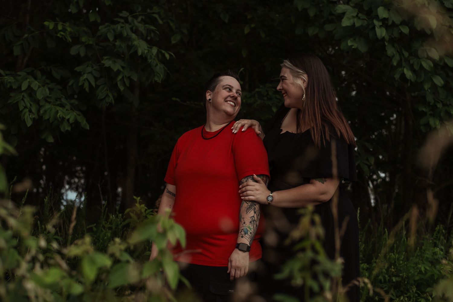 Two people smiling and embracing outdoors among lush green trees, one in a red shirt with tattoos.