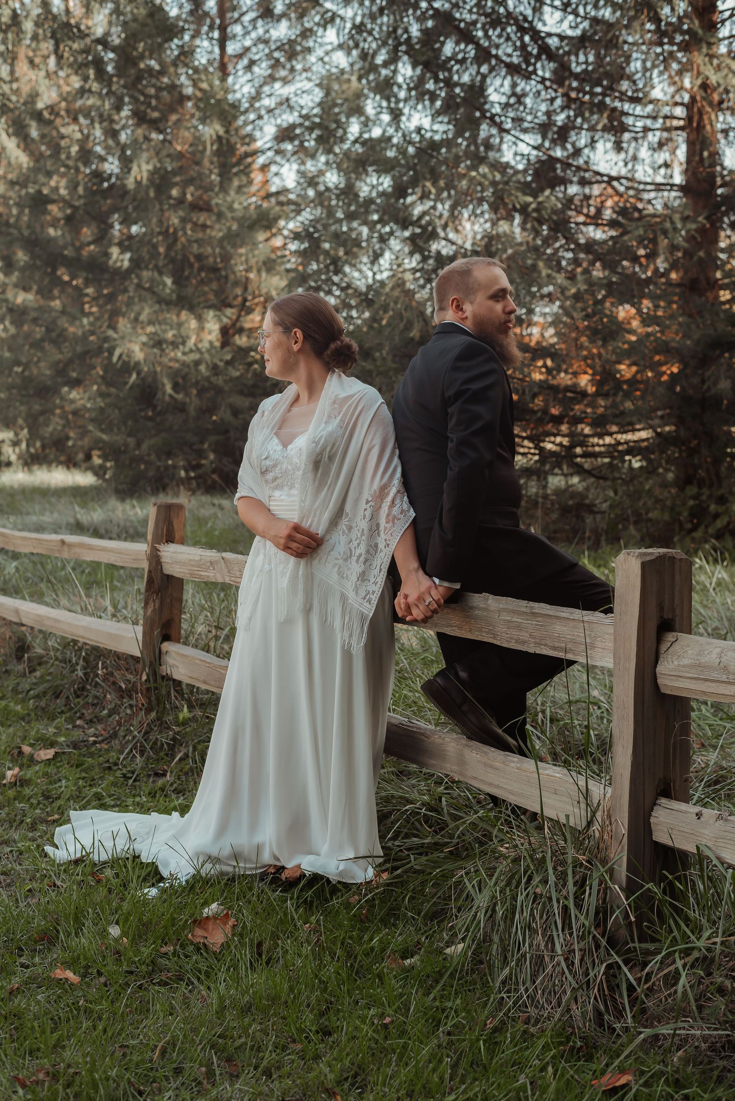 Bride and groom holding hands leaning against rustic wooden fence.