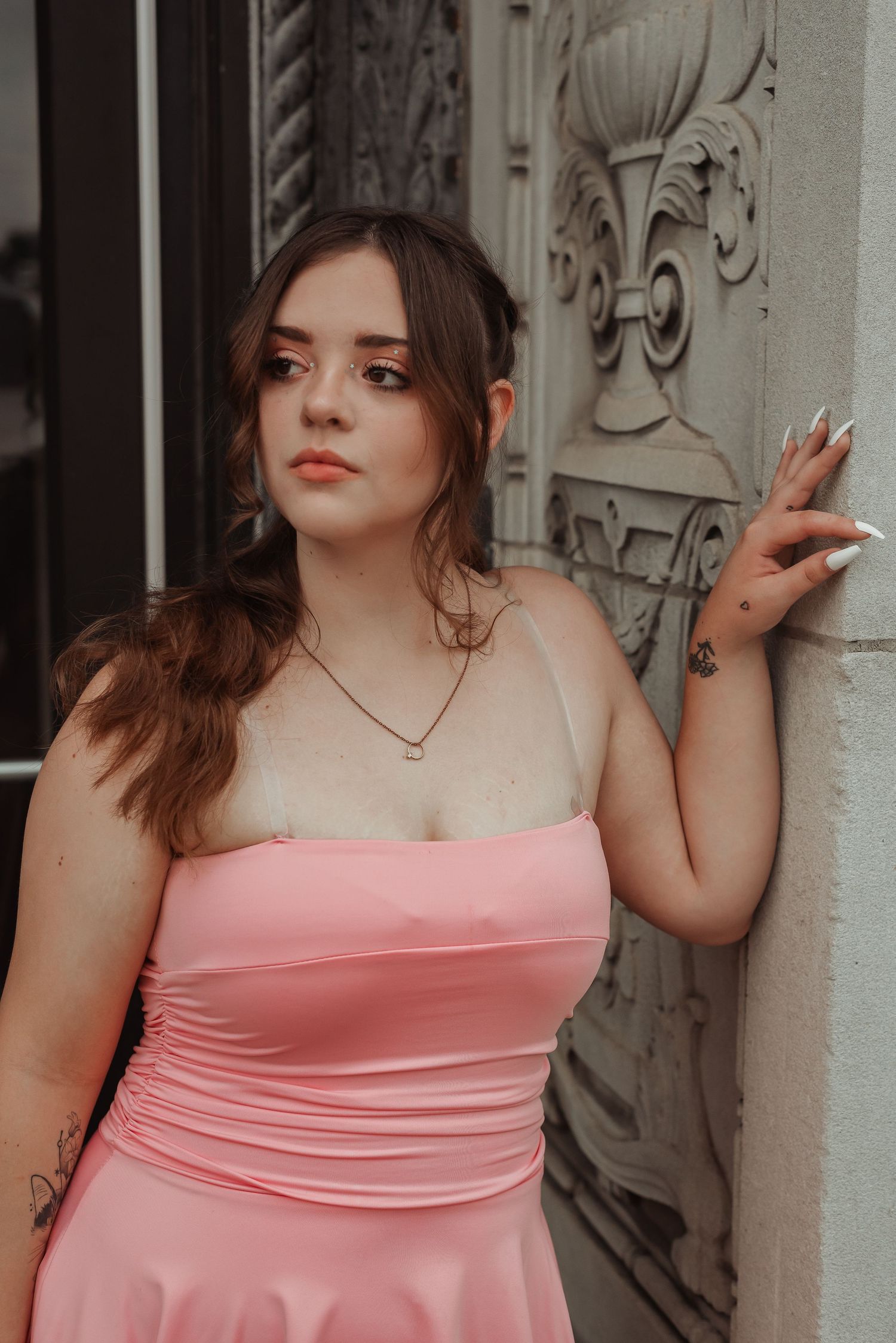 Young woman in pink strapless dress with tattoos leaning against ornate white wall outdoors.
