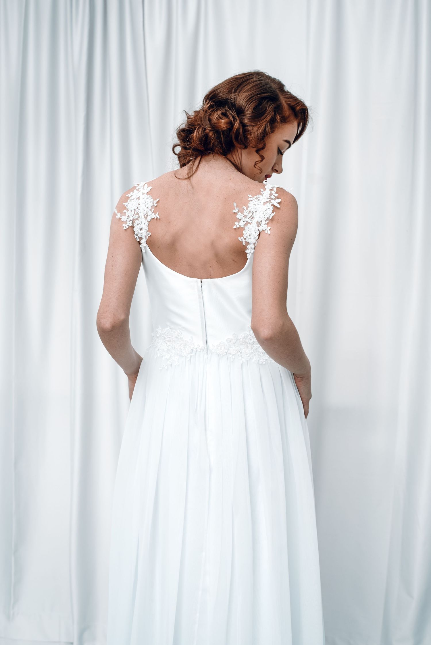 Bride in white A-line gown with floral lace straps standing with back to camera before white curtains.