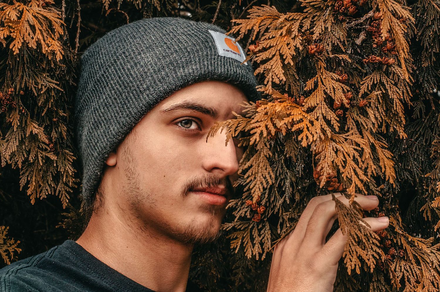 Young man in grey beanie holding autumn foliage near his face outdoors.
