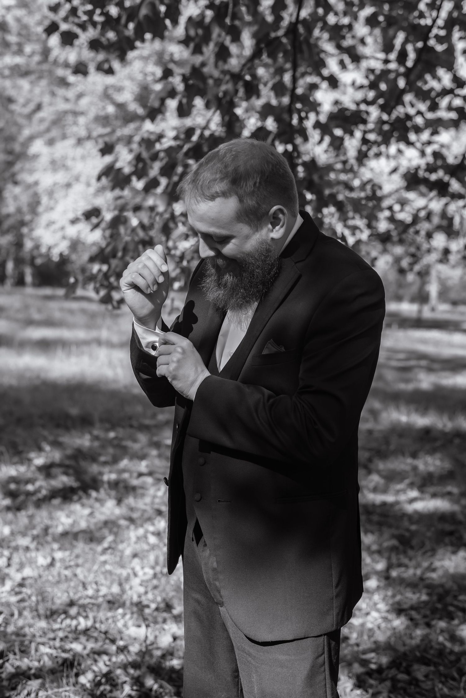 Black and white portrait of a bearded groom in a suit adjusting his cufflinks outdoors in a park.