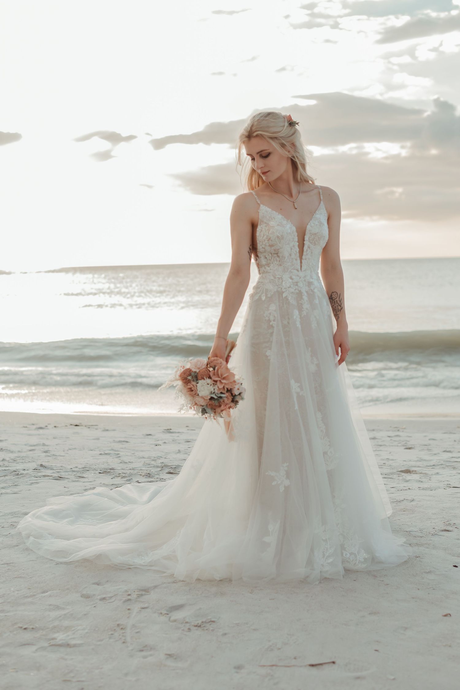 Elegant bride in lace A-line wedding gown holding bouquet on sandy beach at sunset.
