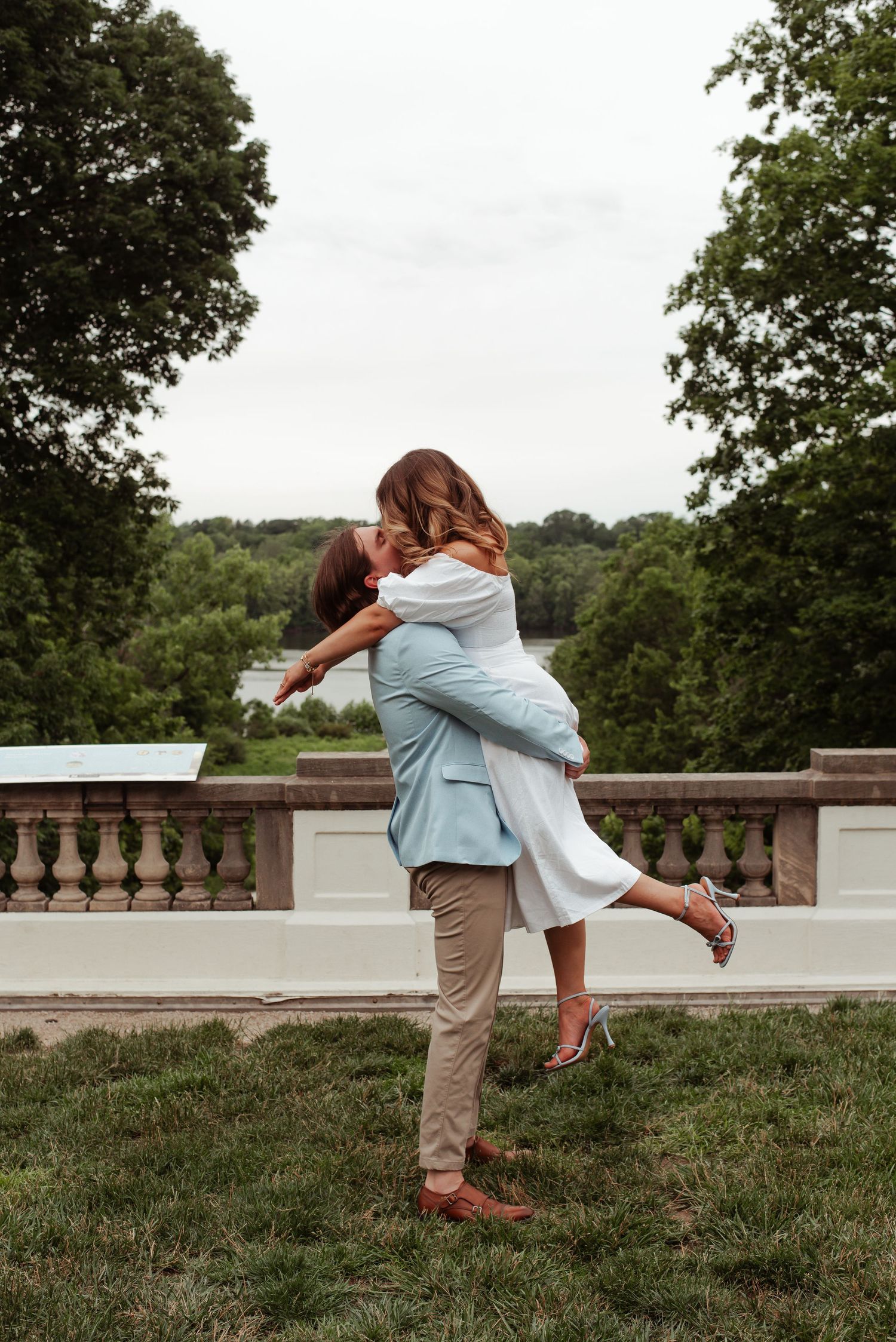 A man lifts a woman in a white dress outdoors, sharing a romantic kiss near a stone balustrade.