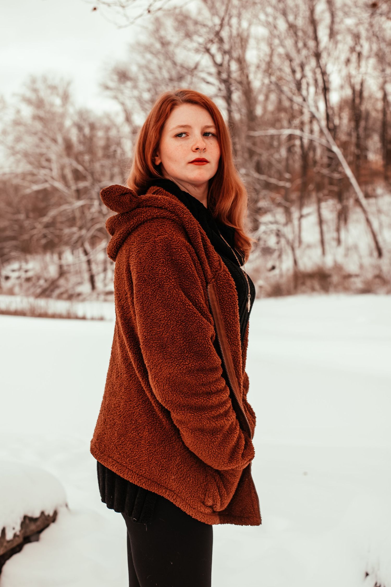 A redhead woman in a brown teddy coat poses outdoors in a snowy winter landscape.
