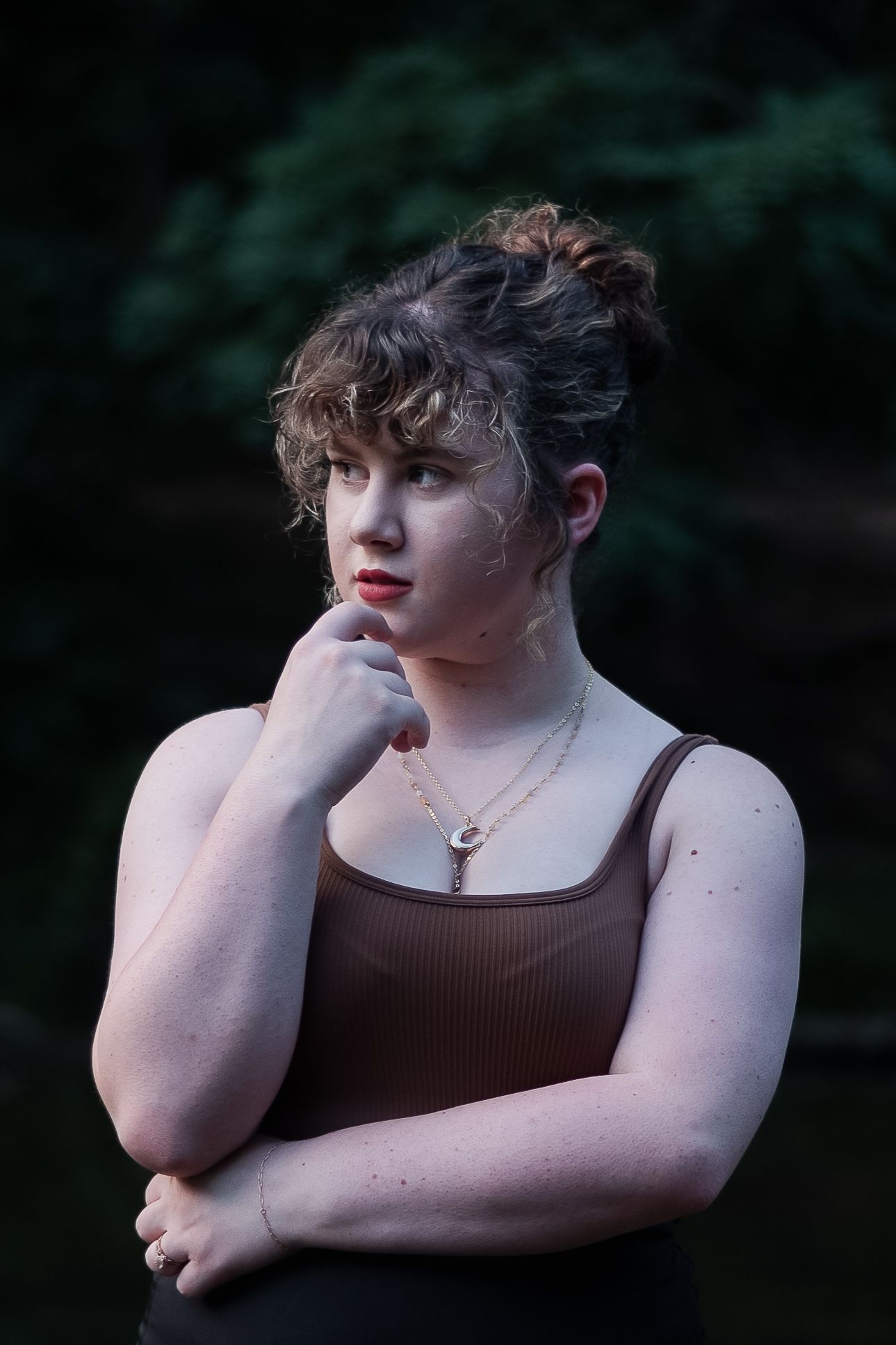 Young woman in brown tank top posing thoughtfully near dark forest backdrop.