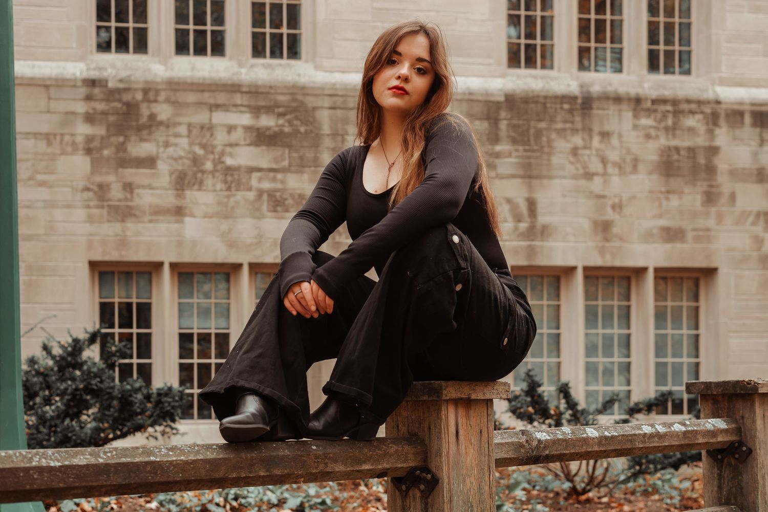 Young woman in all-black outfit sits on wooden fence outside historic stone building.