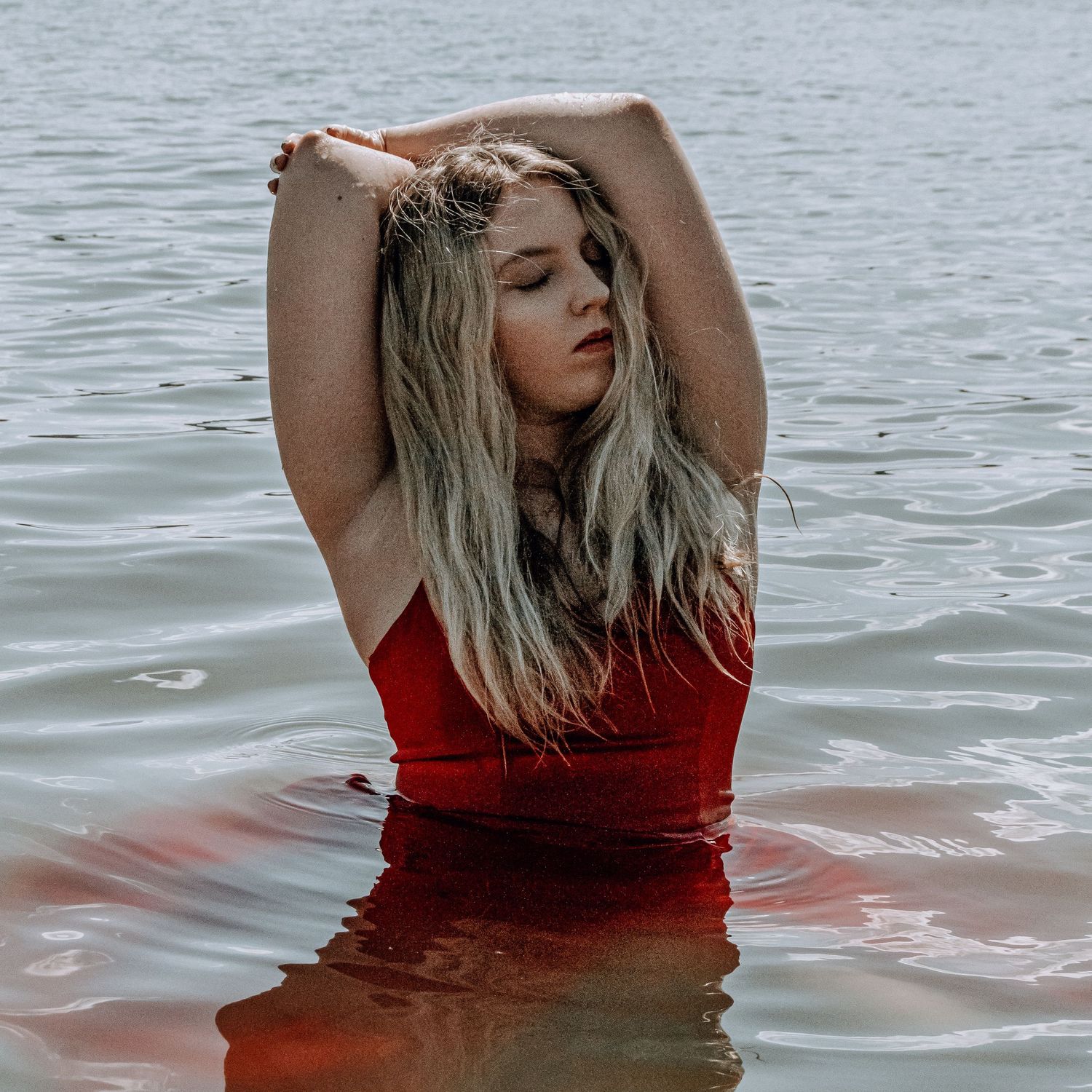 Blonde woman in red swimsuit standing in a lake with arms raised overhead.