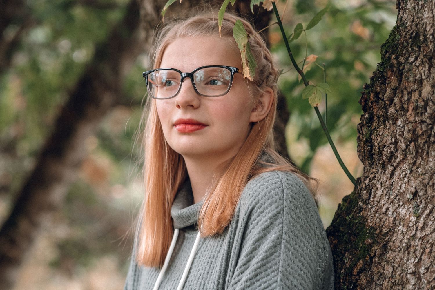 Young blonde woman with red lipstick and glasses leaning against a tree outdoors.