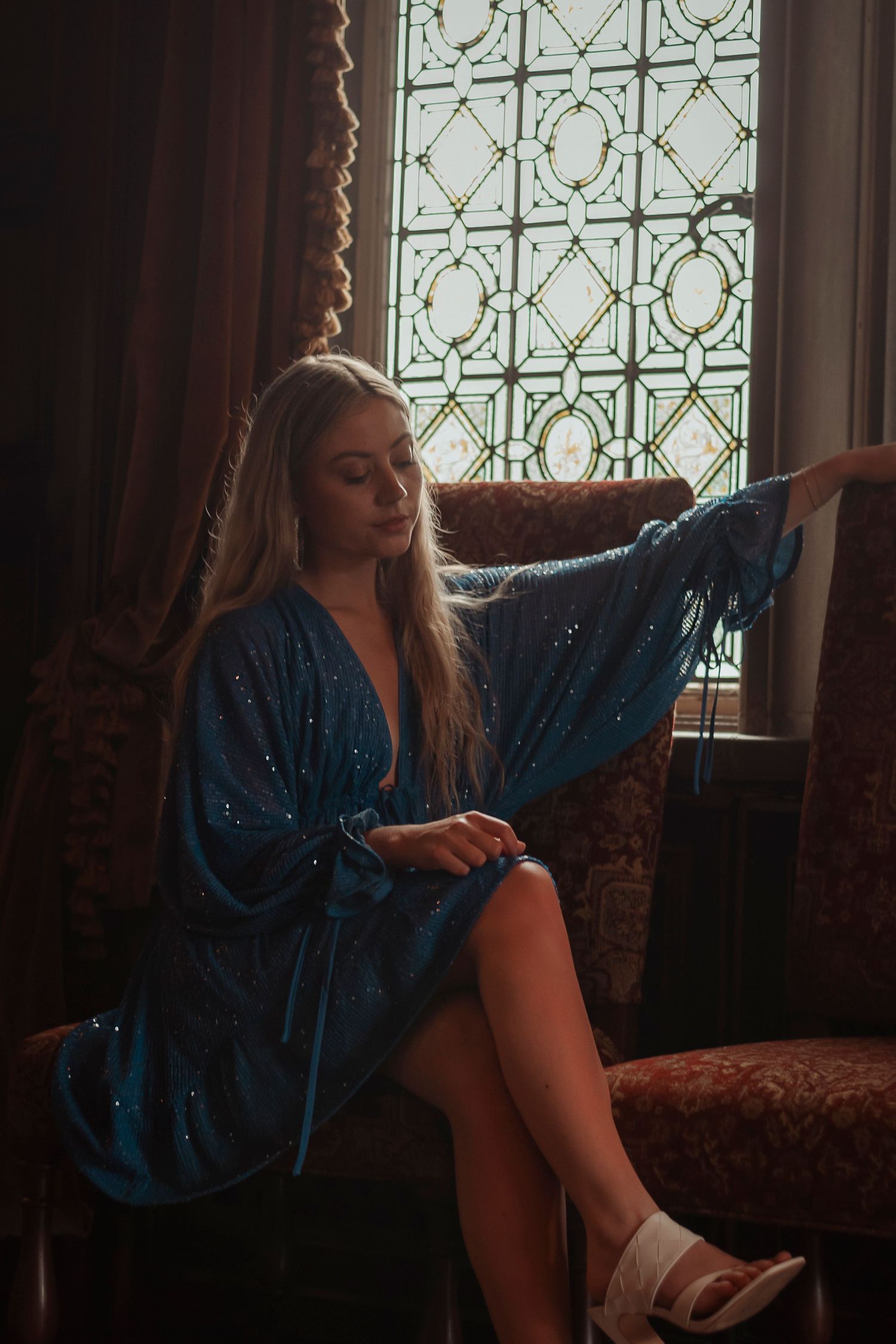 Blonde woman in blue sequin kimono robe seated by stained glass window in vintage armchair.