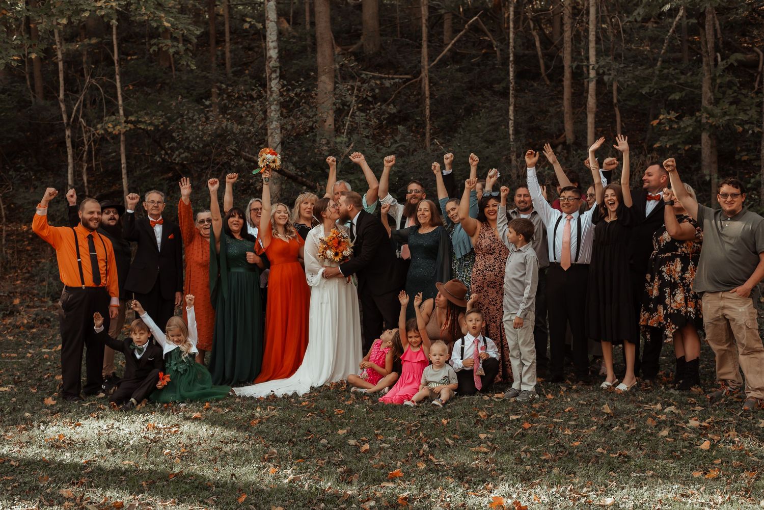 Large wedding party celebrating outdoors in forest setting with bride and groom centered among guests raising hands.