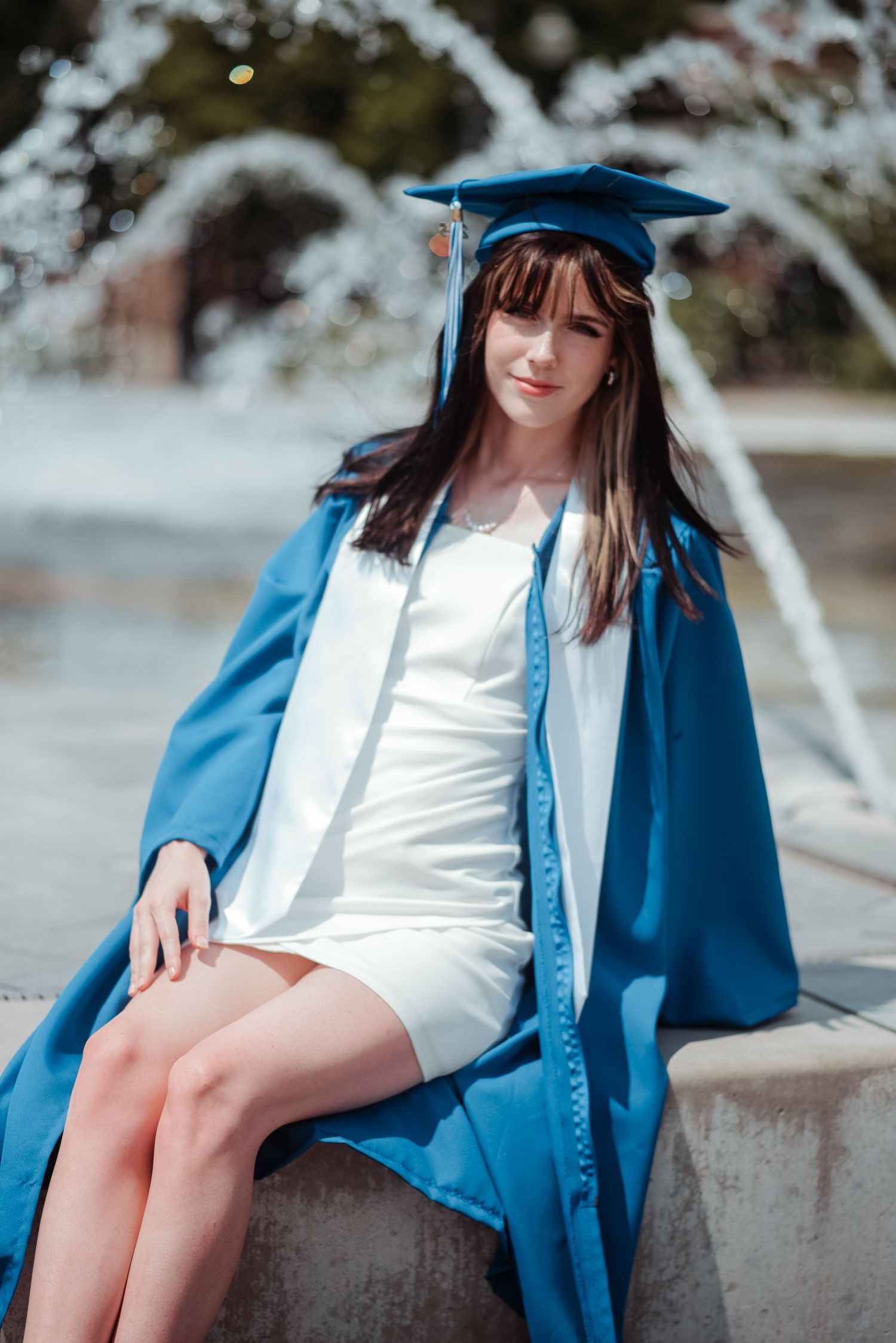 Female graduate in blue cap and gown over white dress sitting by outdoor fountain in sunny campus setting.