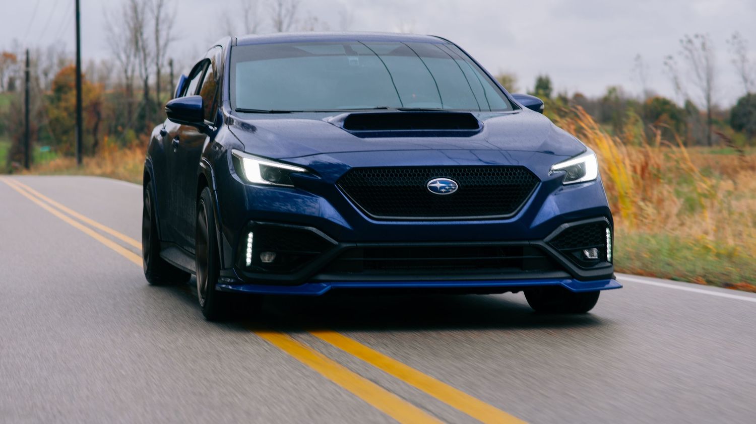 Dark blue Subaru WRX sedan driving on a road surrounded by autumn foliage.