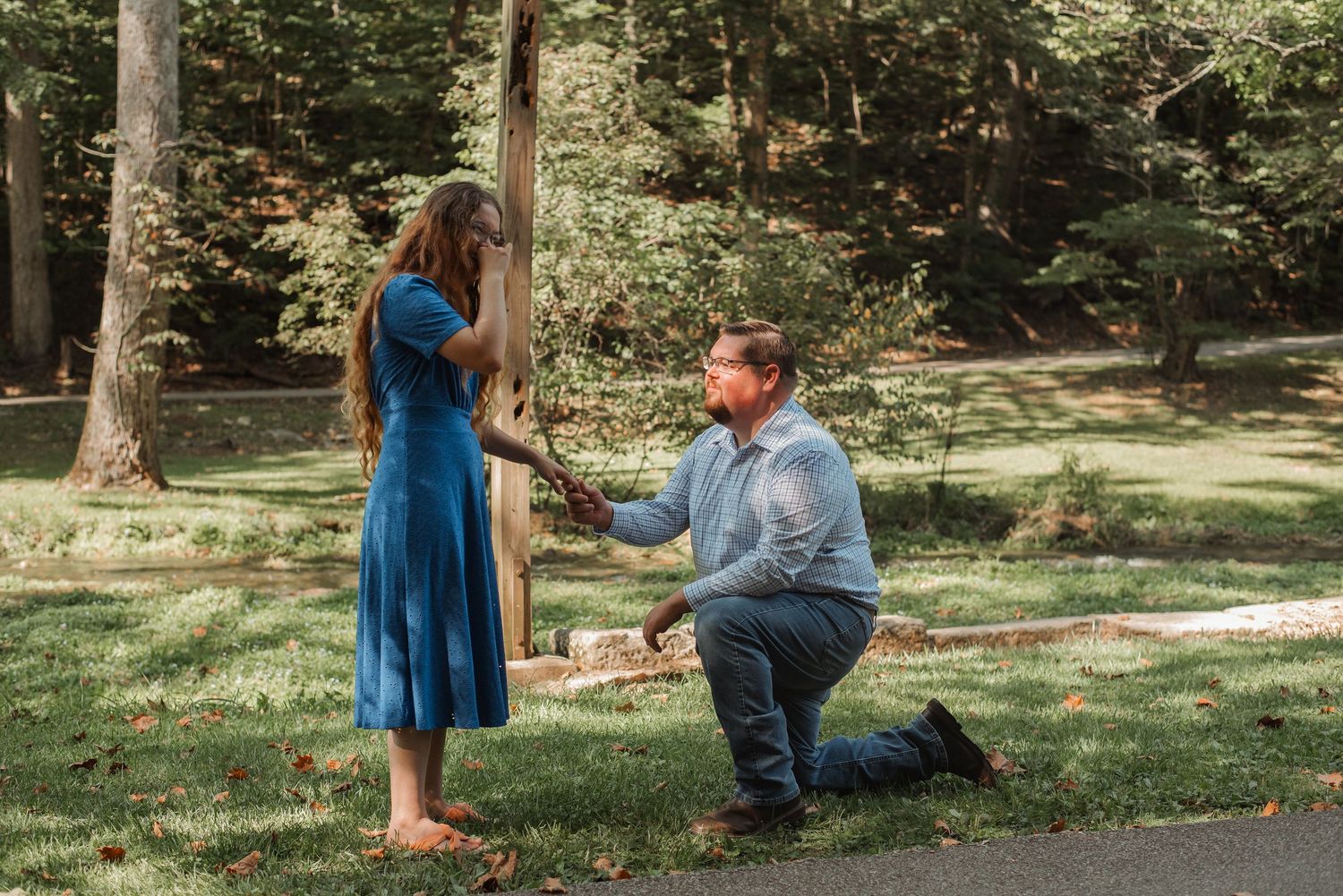Man proposing on one knee to woman in blue dress outdoors in lush green park setting.