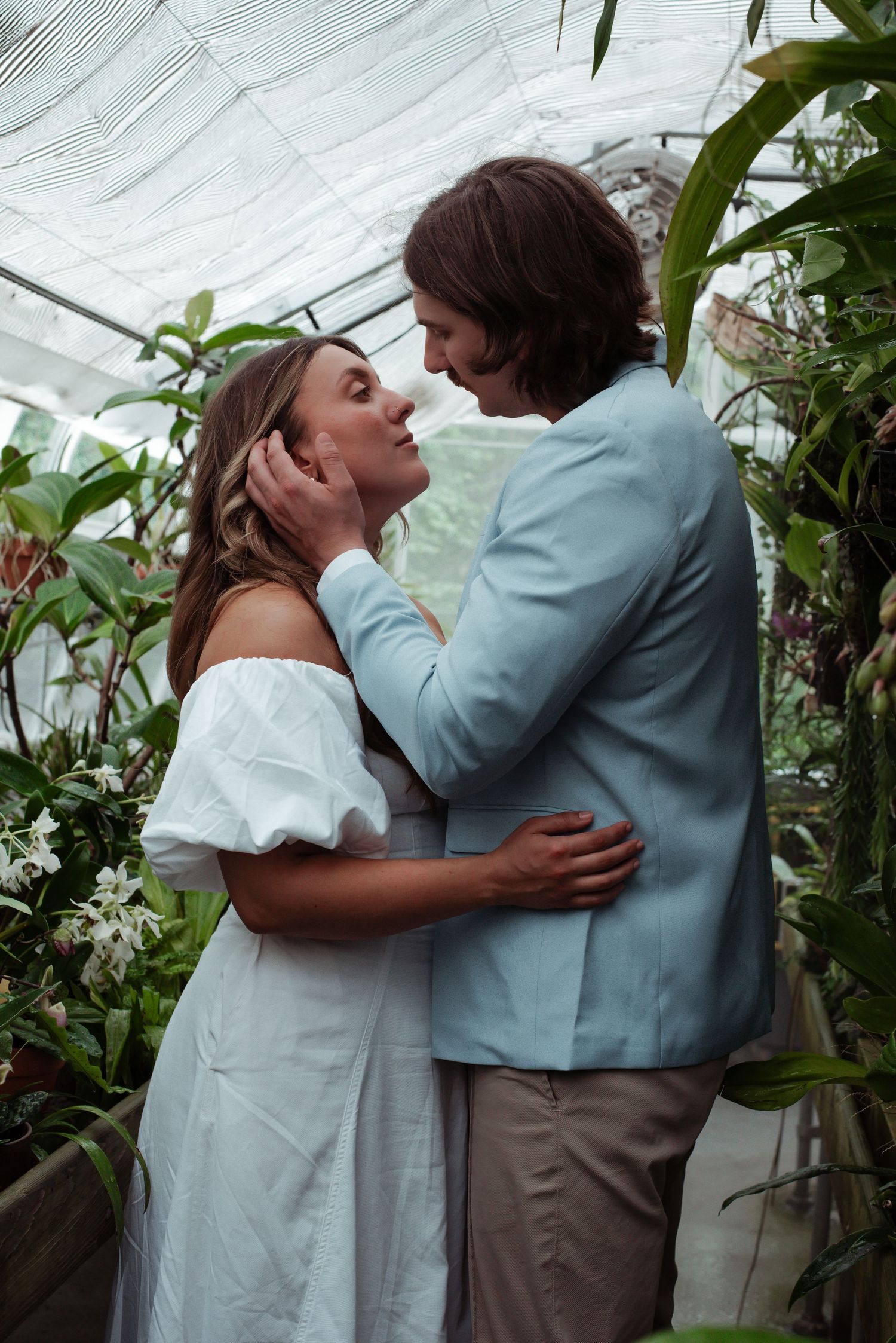 Couple sharing a romantic moment in a lush greenhouse, woman in white dress, man in light blue suit.