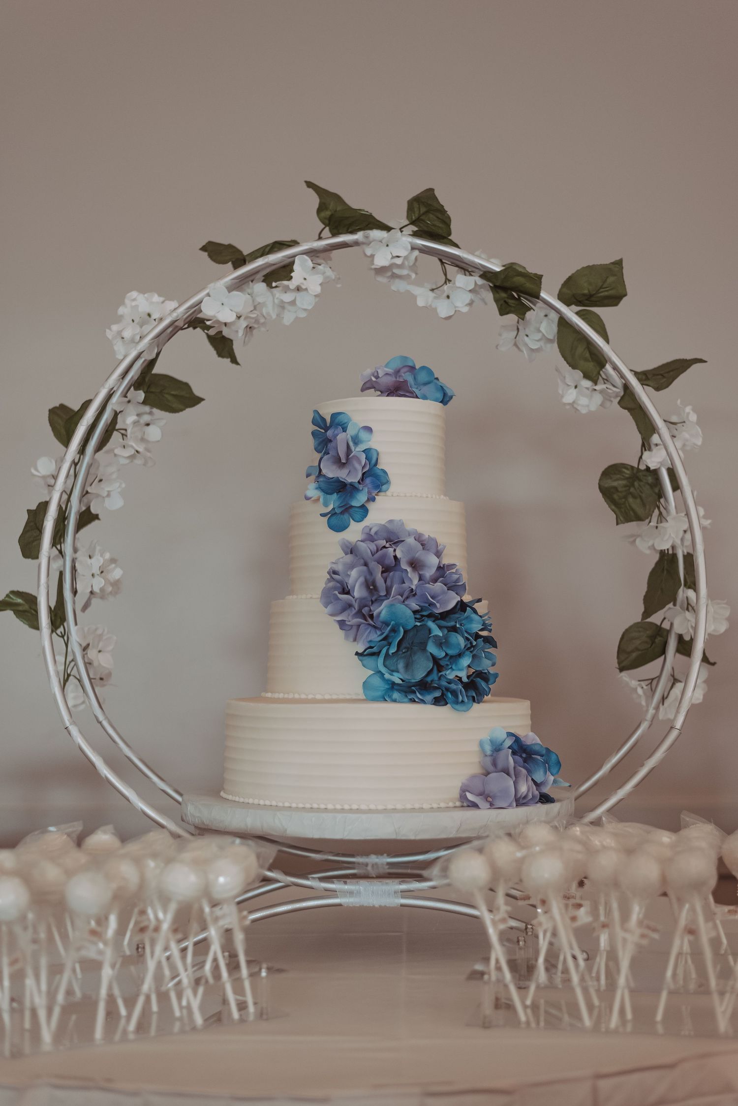 Elegant four-tier white wedding cake decorated with blue and purple hydrangeas displayed in circular floral hoop stand.