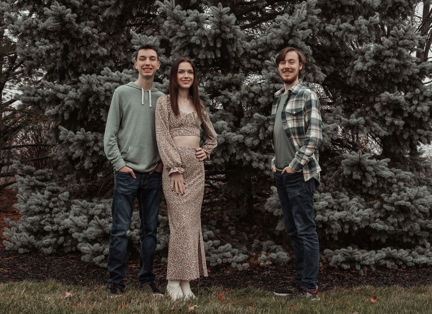 Three siblings pose together outdoors in front of tall evergreen trees for a family portrait.