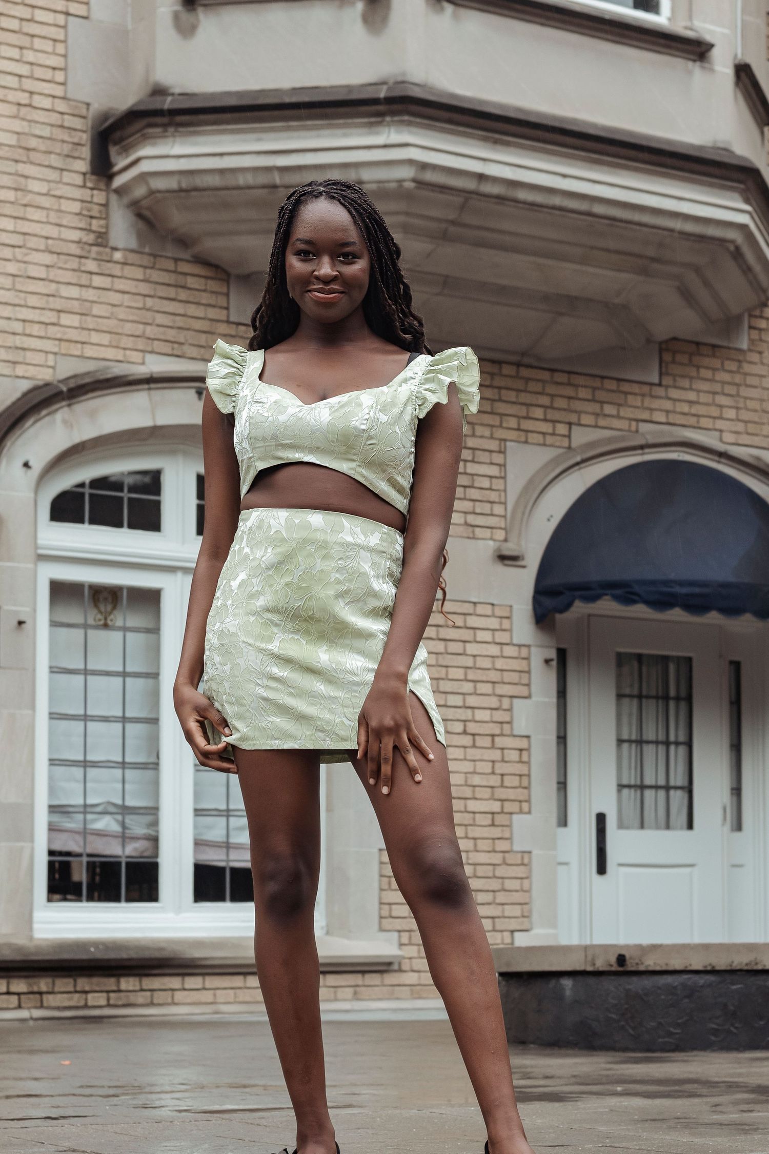 Smiling young woman in mint green two-piece outfit standing in front of classic brick building.