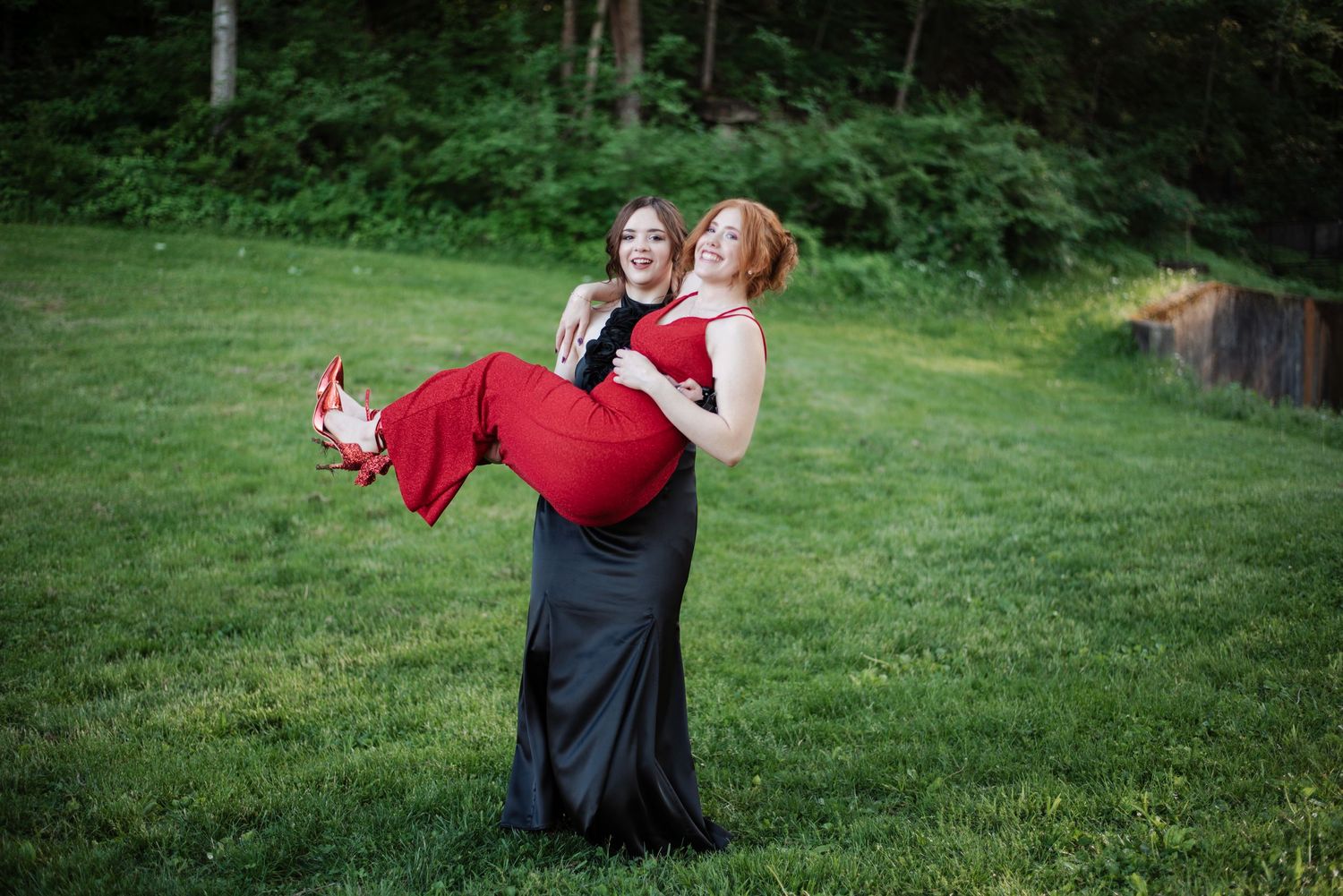 Two prom girls pose outdoors, one in black gown lifting friend in red dress on green lawn.
