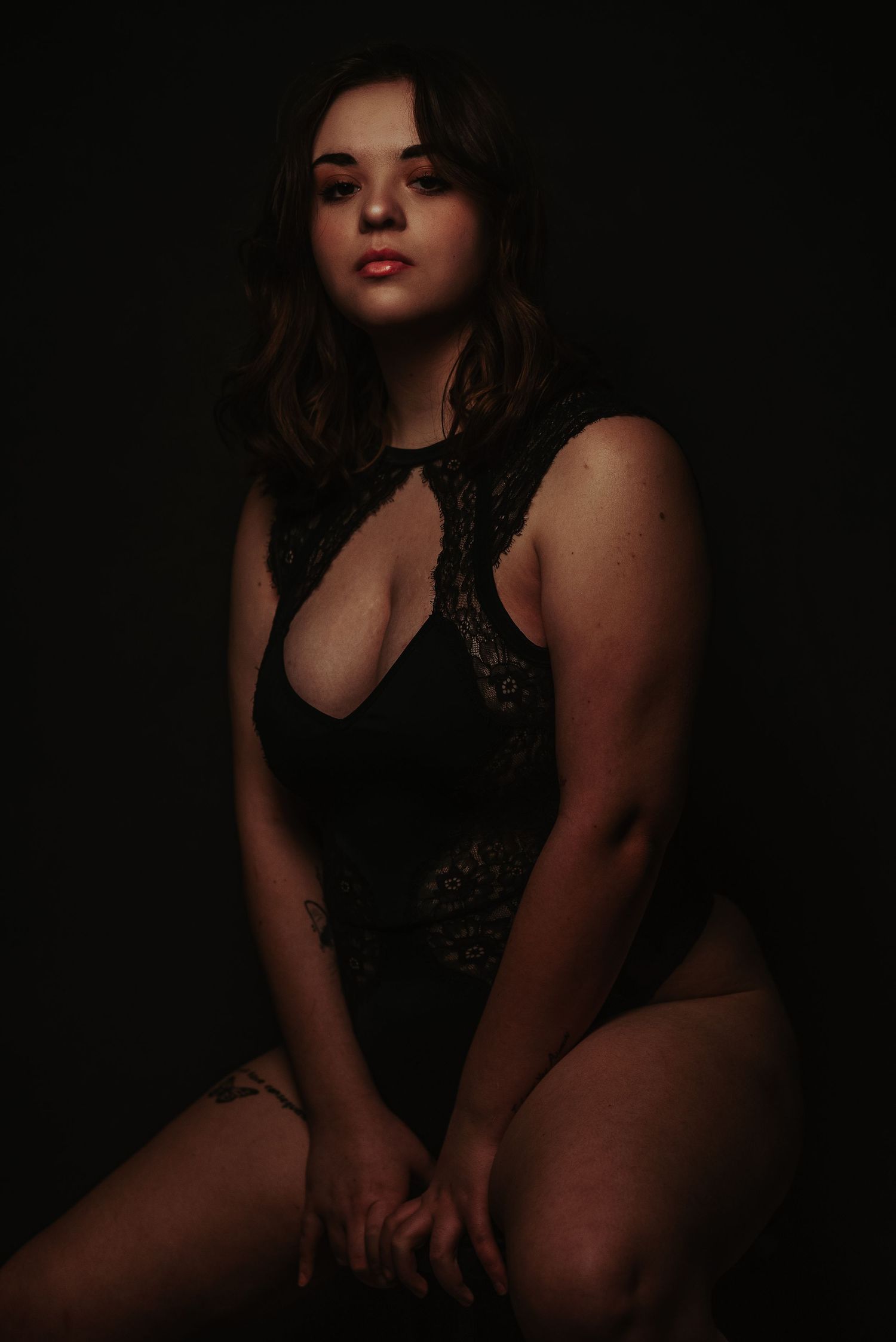 Woman in black lace bodysuit posing confidently in dark studio setting.