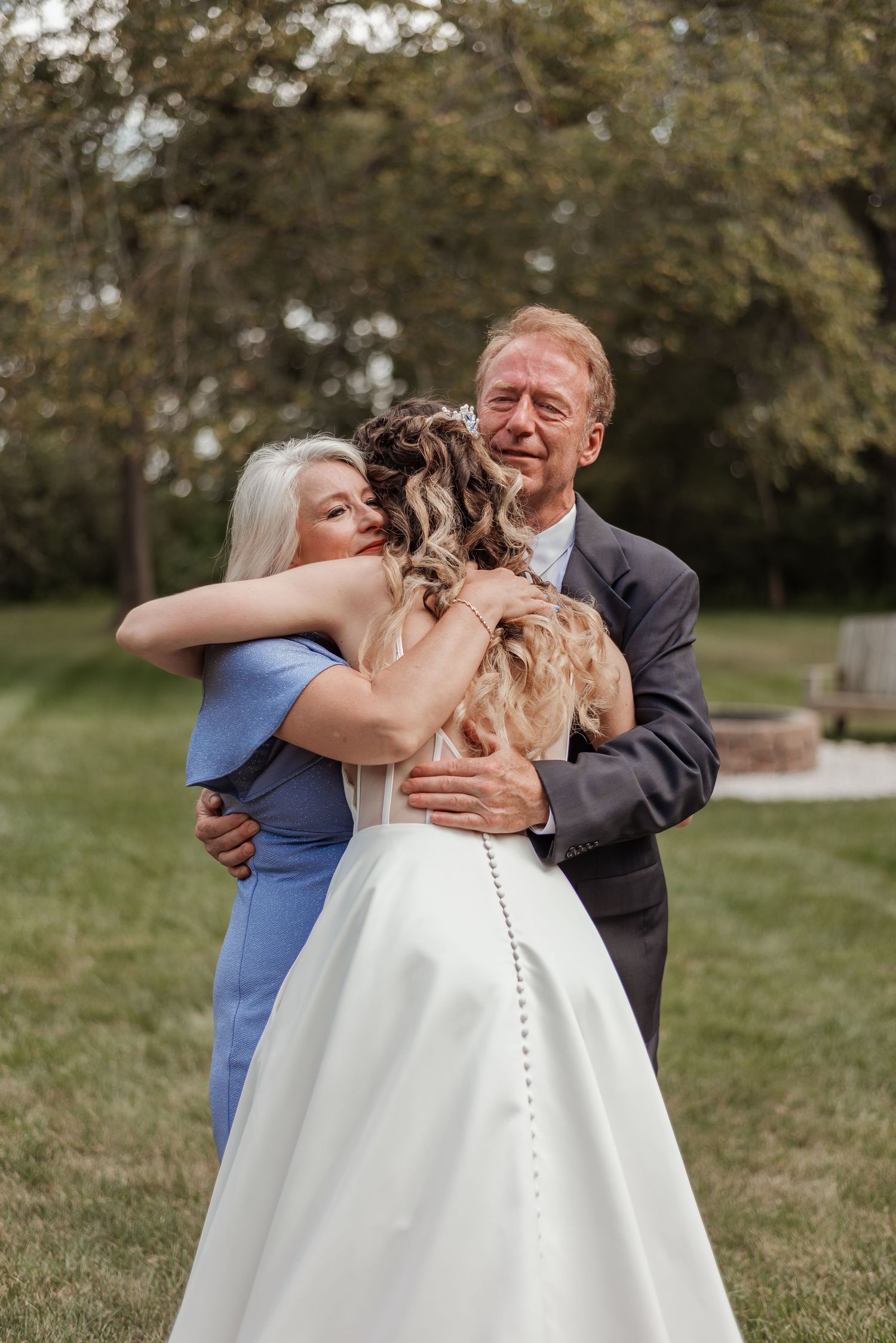 Emotional bride embracing her parents in a heartfelt group hug outdoors before wedding ceremony.