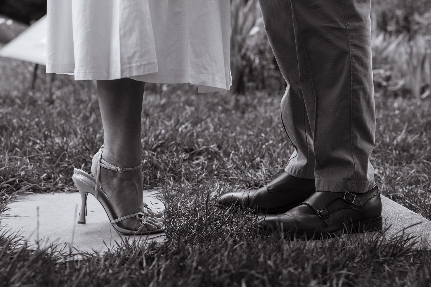 Black and white photo of a bride in heels and groom in dress shoes standing on a stone at an outdoor wedding.