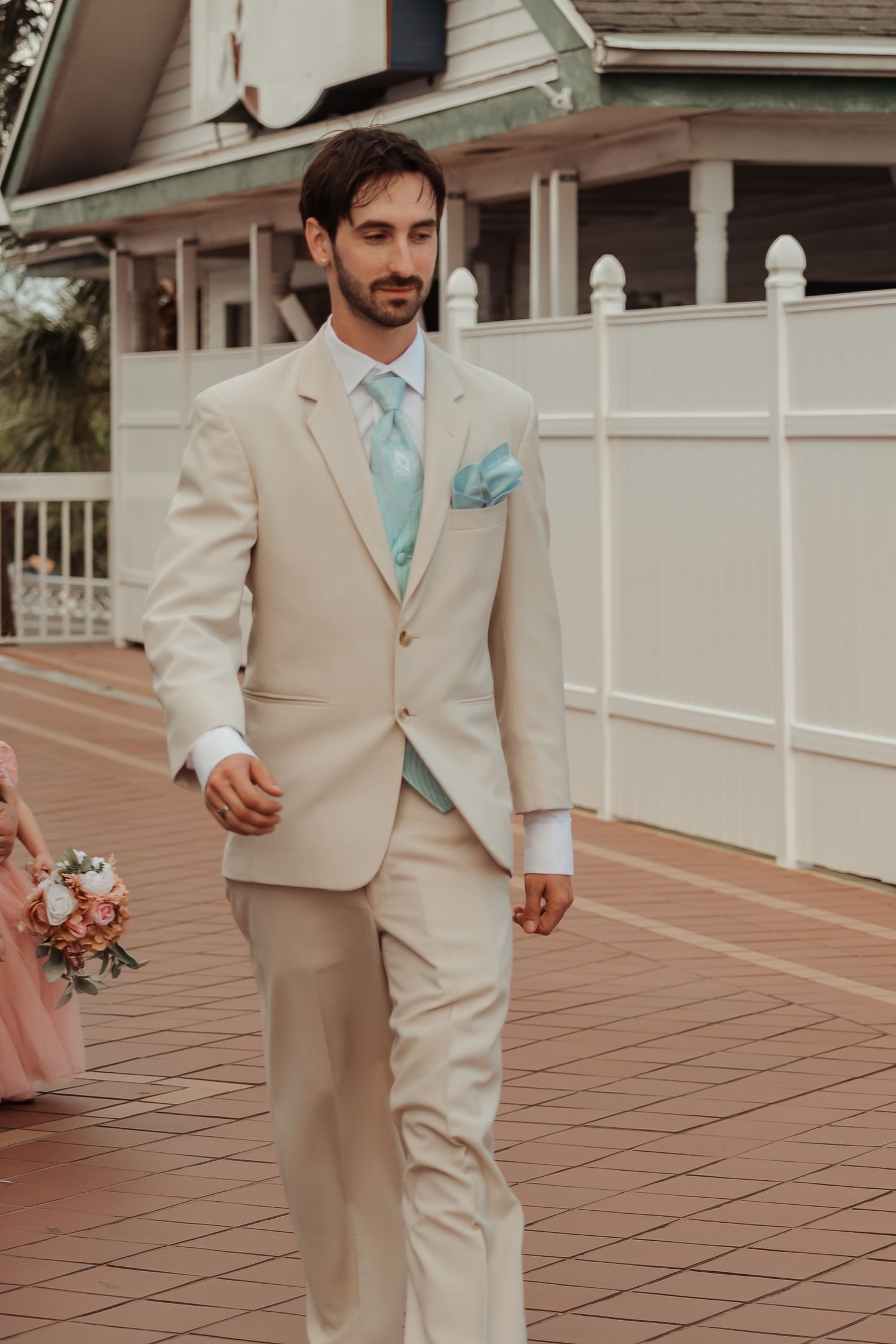 Handsome groom in beige suit with mint tie and pocket square walking at outdoor wedding venue.