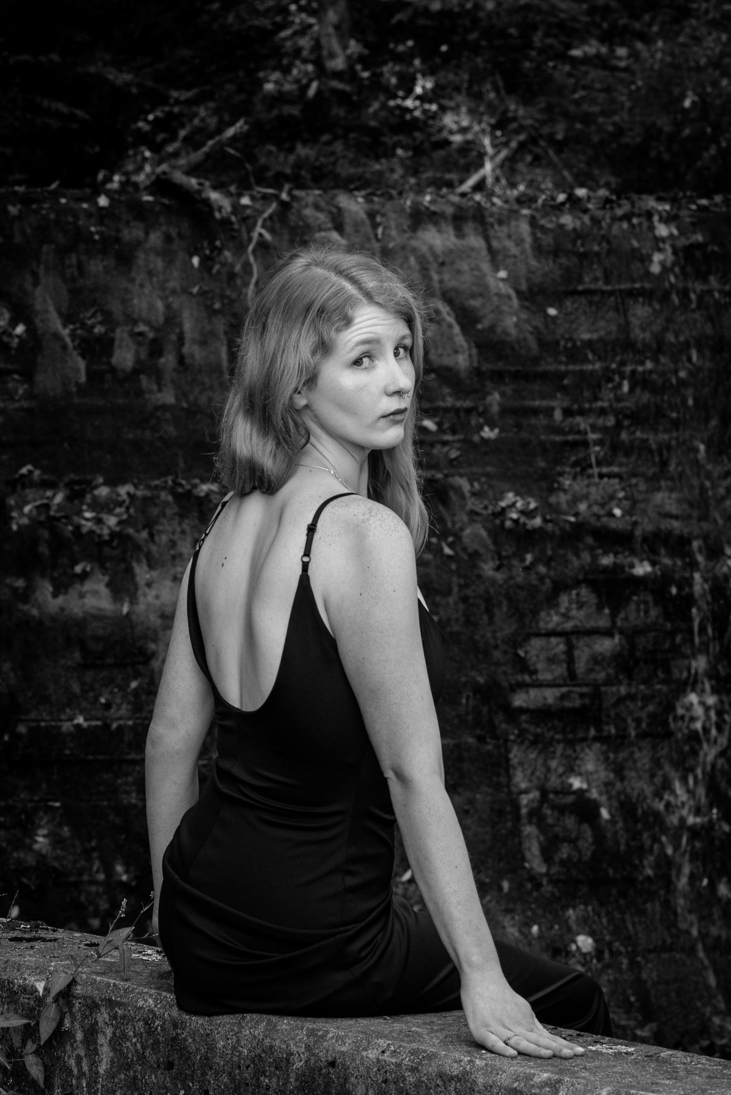 Black and white portrait of woman in black backless dress sitting against stone wall outdoors.