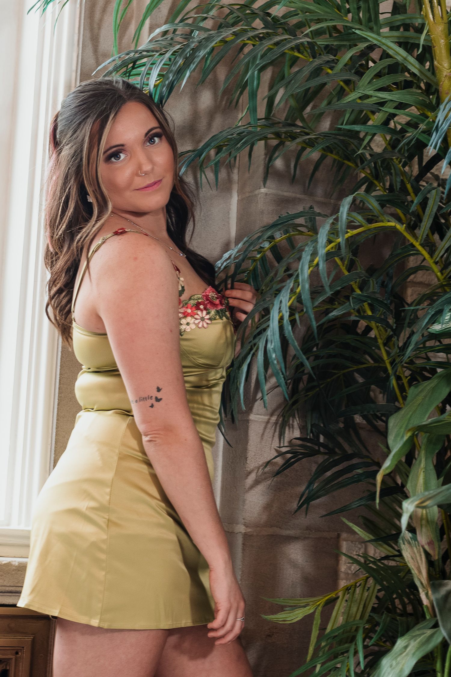Brunette woman in a green satin mini dress posing beside a large tropical plant near a window.