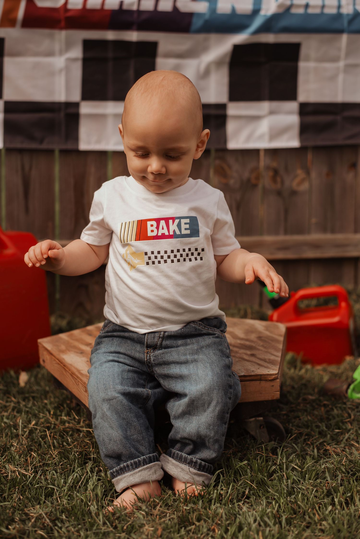 A bald baby in a BAKE shirt and jeans sits on a wooden board at a racing-themed birthday party.
