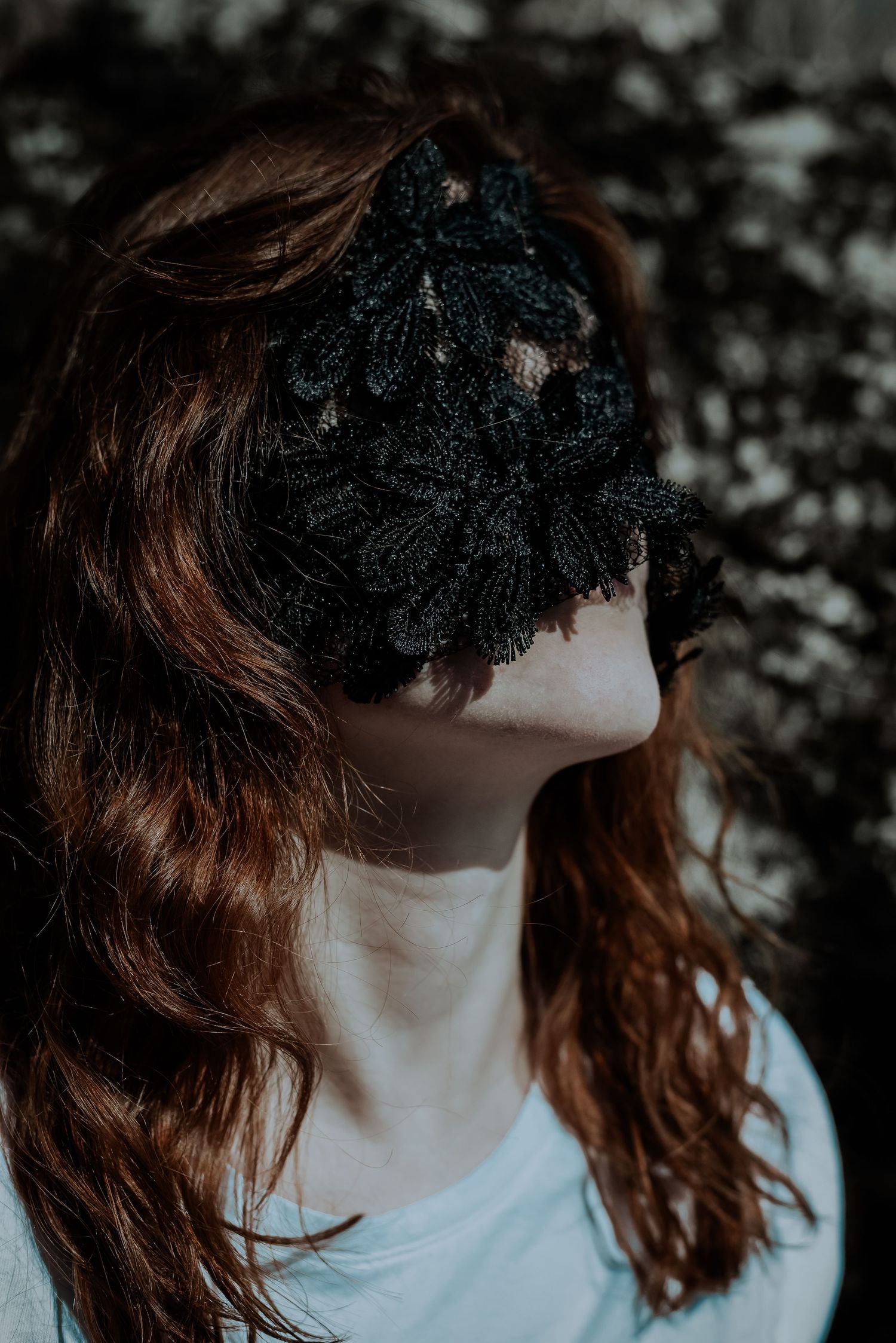 Redhead woman wearing black floral lace mask covering eyes in dark outdoor setting.