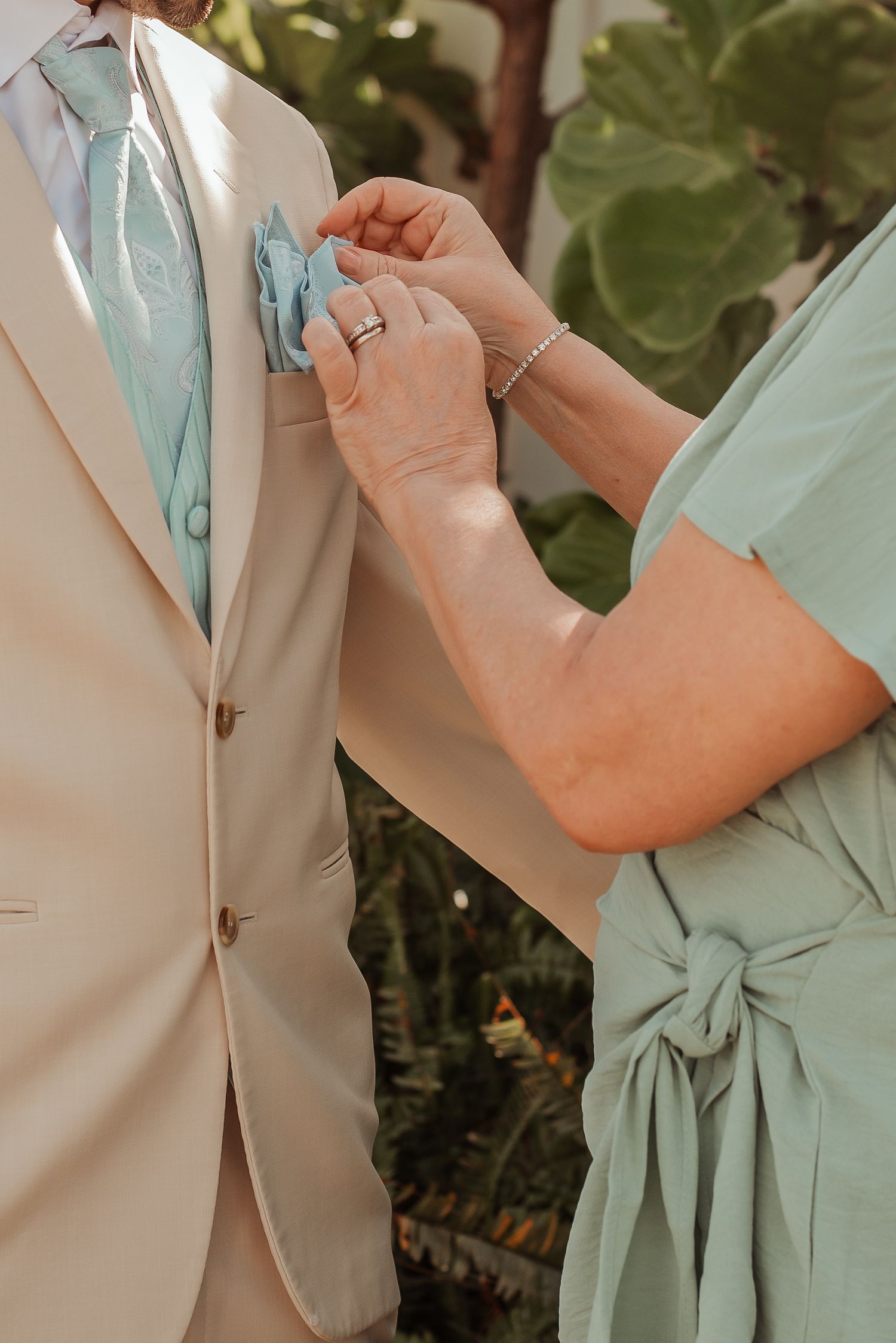 Mother of groom adjusting mint pocket square on grooms tan suit before wedding ceremony.