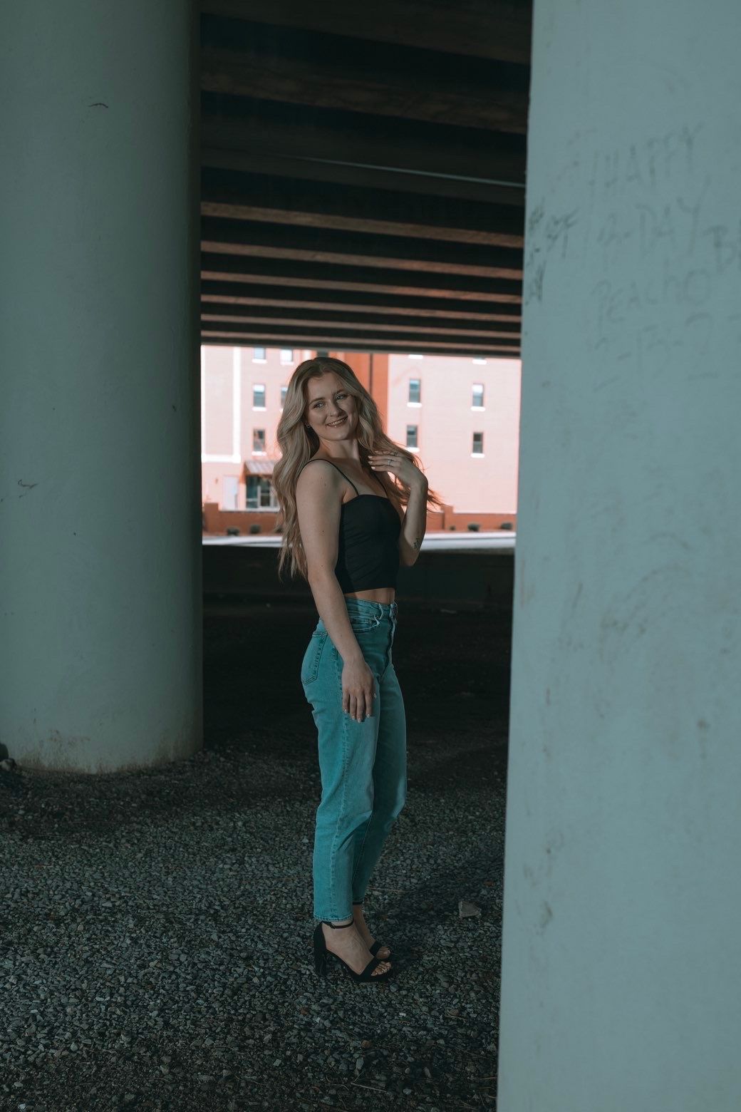 Blonde woman in black crop top and jeans posing under an urban bridge.