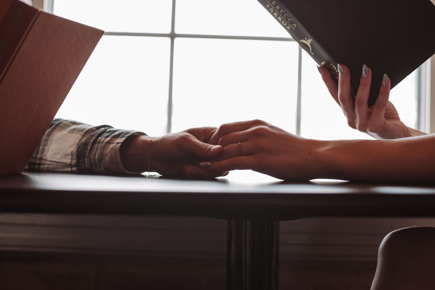 Two people holding hands across a wooden table near a sunlit window indoors.
