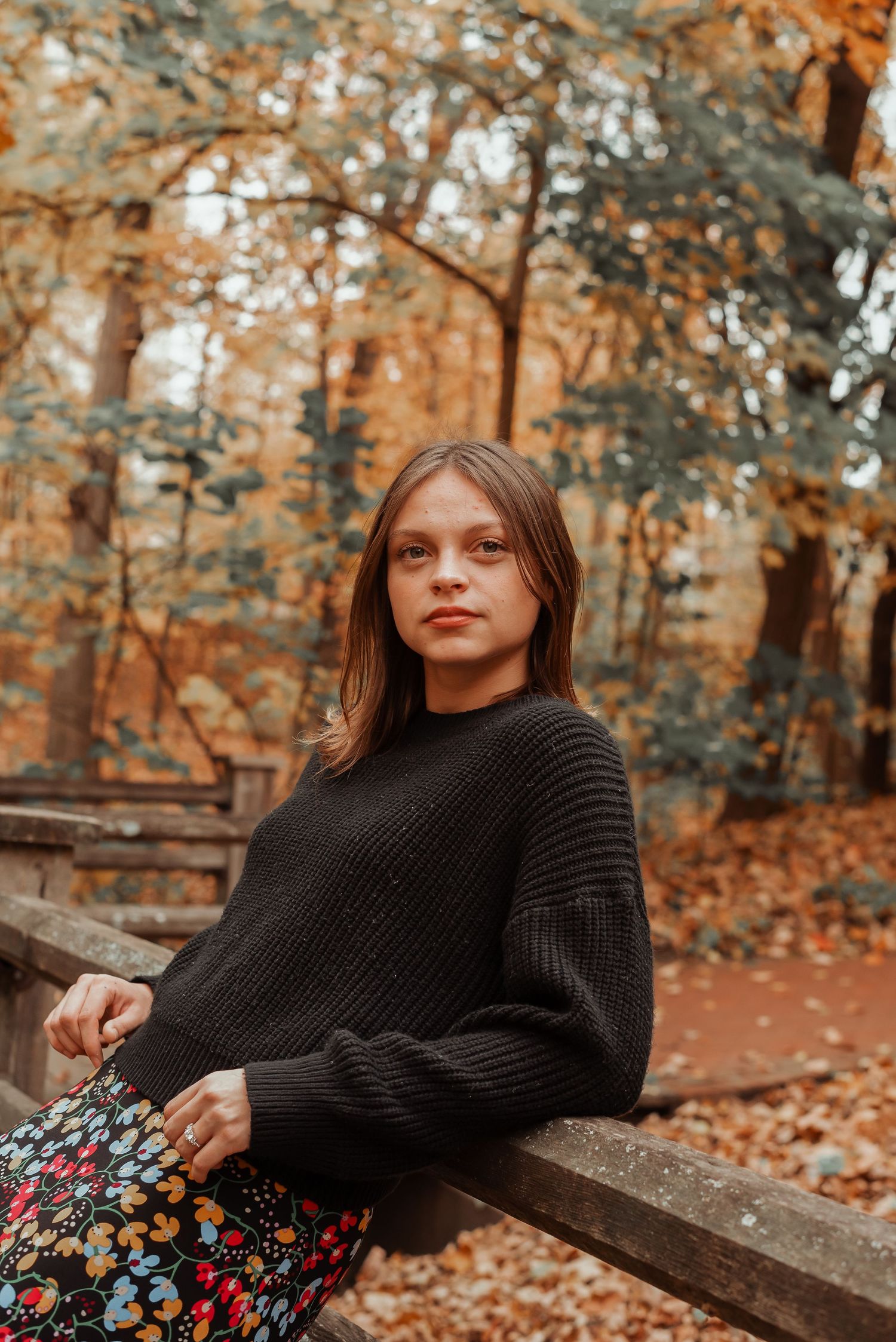 Young woman in dark knit sweater and floral skirt sits on wooden fence in autumn forest.