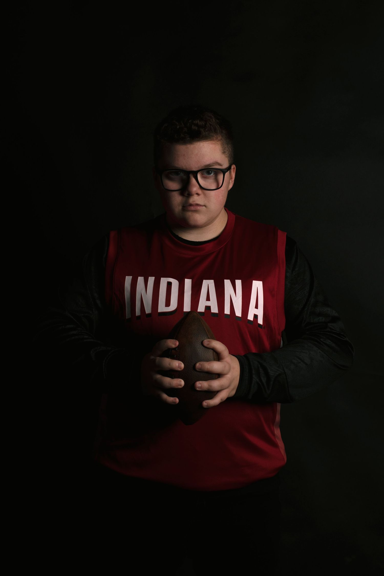 Teen boy in Indiana jersey holding football against dark background.