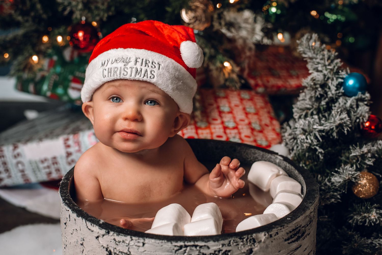 Sweet baby wearing My First Christmas Santa hat poses in marshmallow-filled barrel by decorated tree.