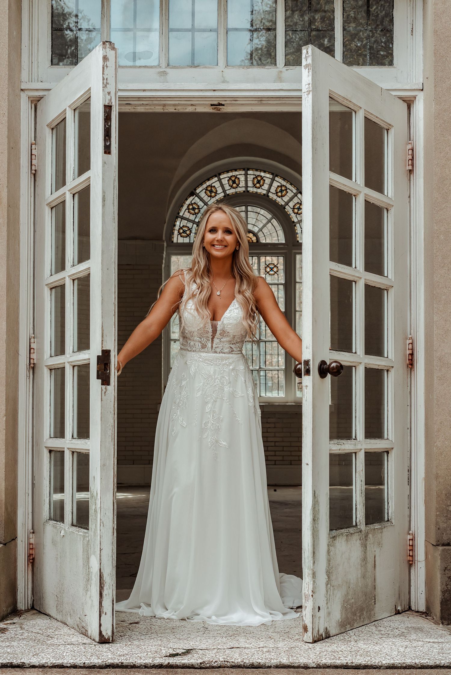 Radiant bride in deep V-neck lace wedding gown stands in open white French doors of historic venue.