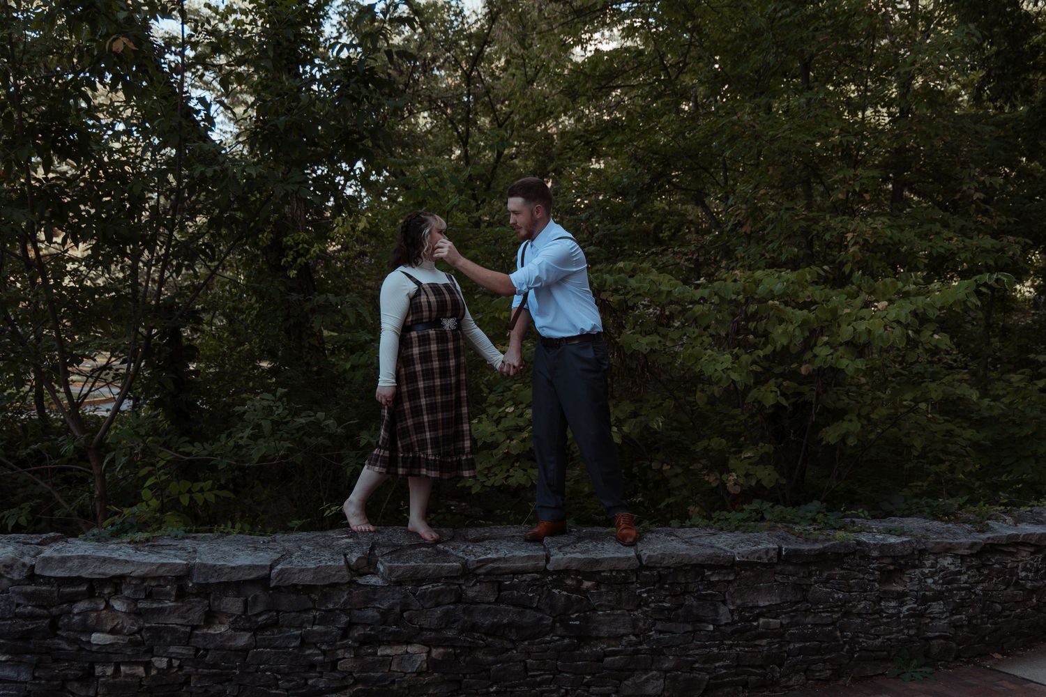 A couple holds hands and dances on a stone wall surrounded by lush green trees at dusk.