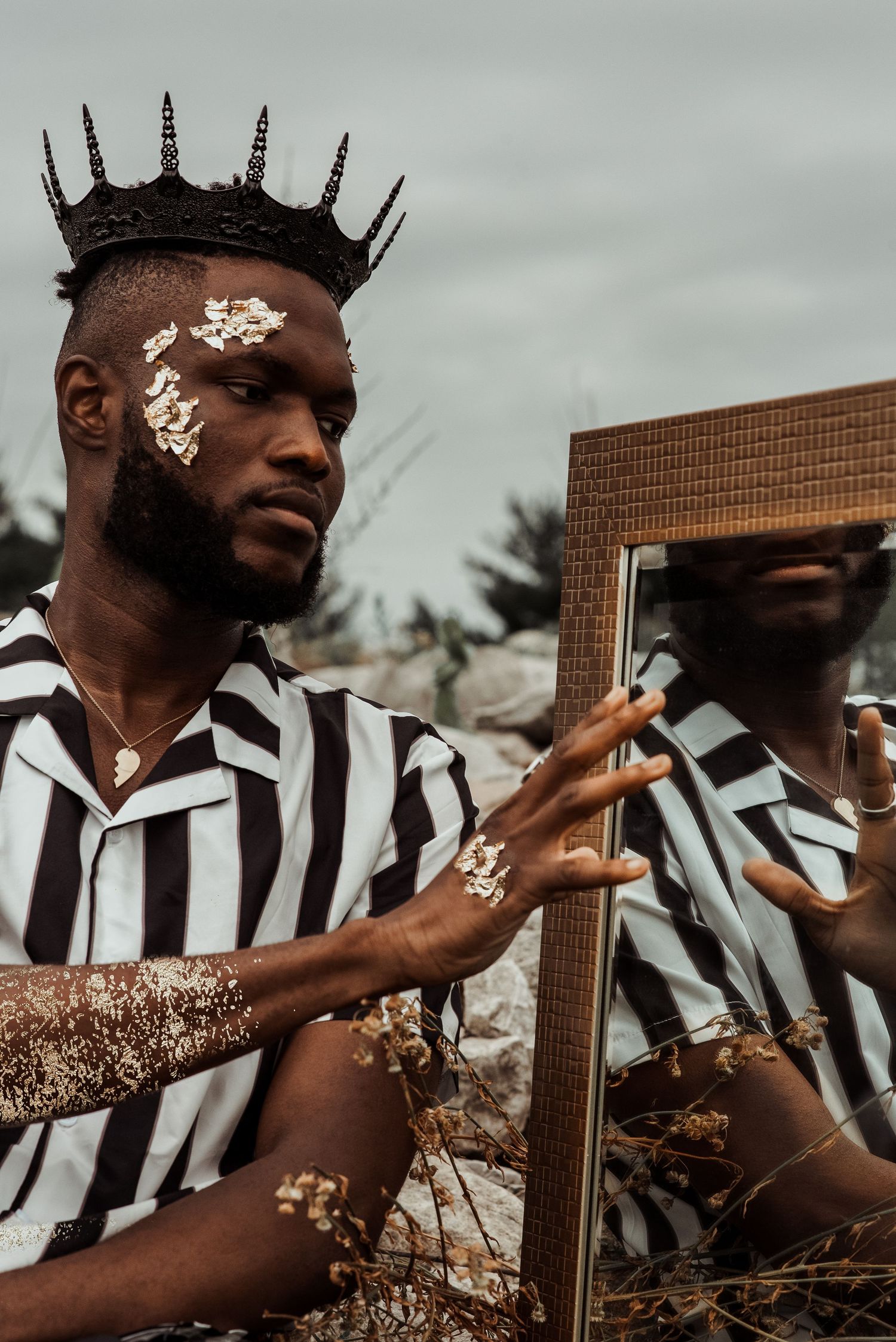 A man wearing a black crown and striped shirt poses beside a mirror with flowers on his face.