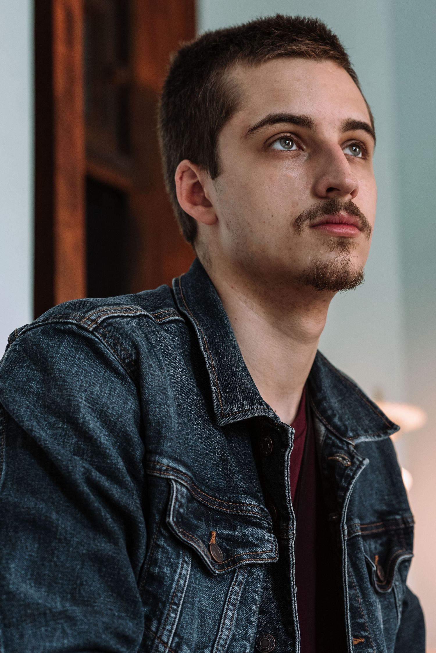 Young man with mustache in denim jacket looking upward indoors.