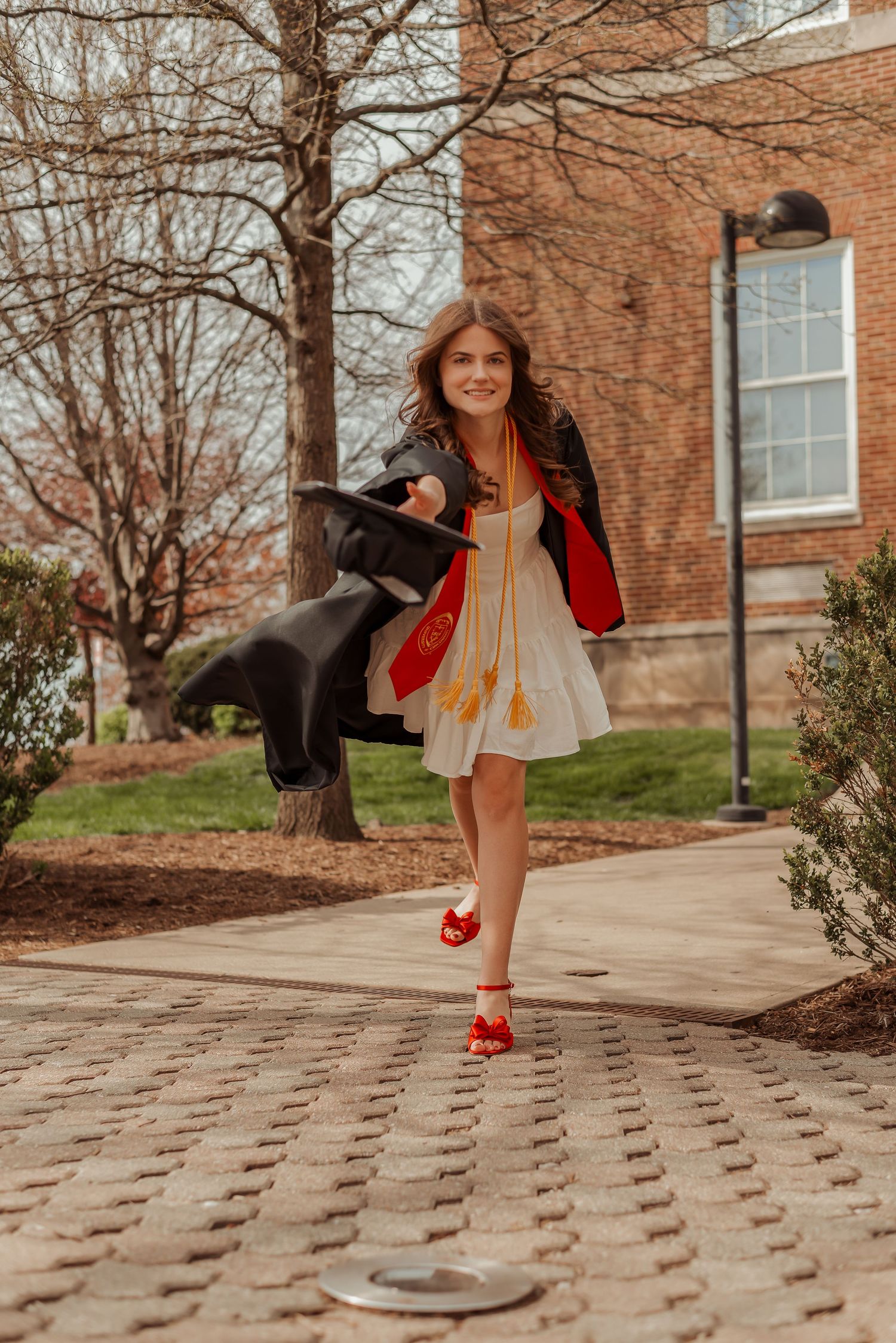 Joyful graduate in white dress with red hood and black gown walks on brick campus path at University of Cincinnati.