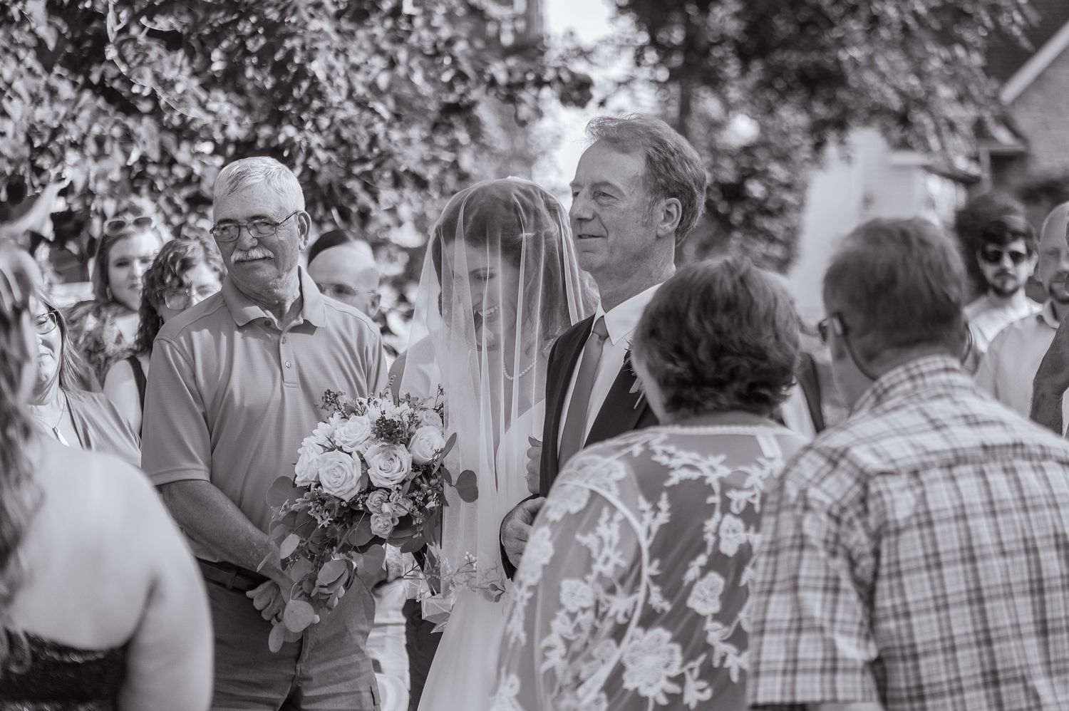 Black and white photo of veiled bride with bouquet walking with father through wedding guests.