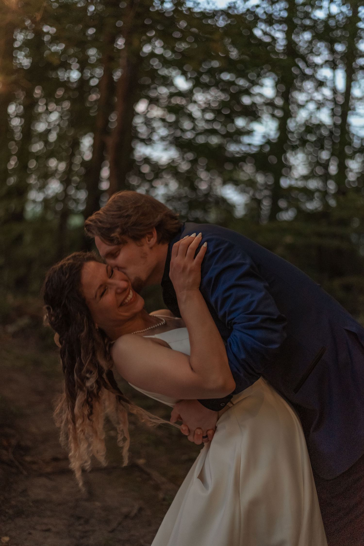 Bride and groom sharing tender embrace in forest at golden hour, bride smiling joyfully.