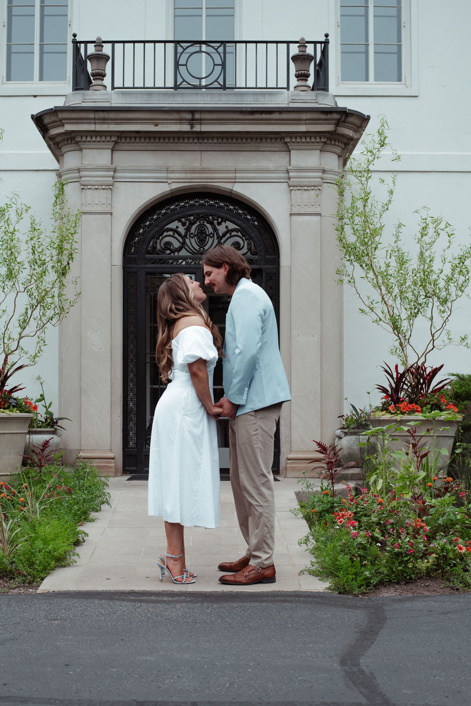 Couple sharing a romantic kiss in white dress and blue suit before elegant mansion entrance with lush gardens.