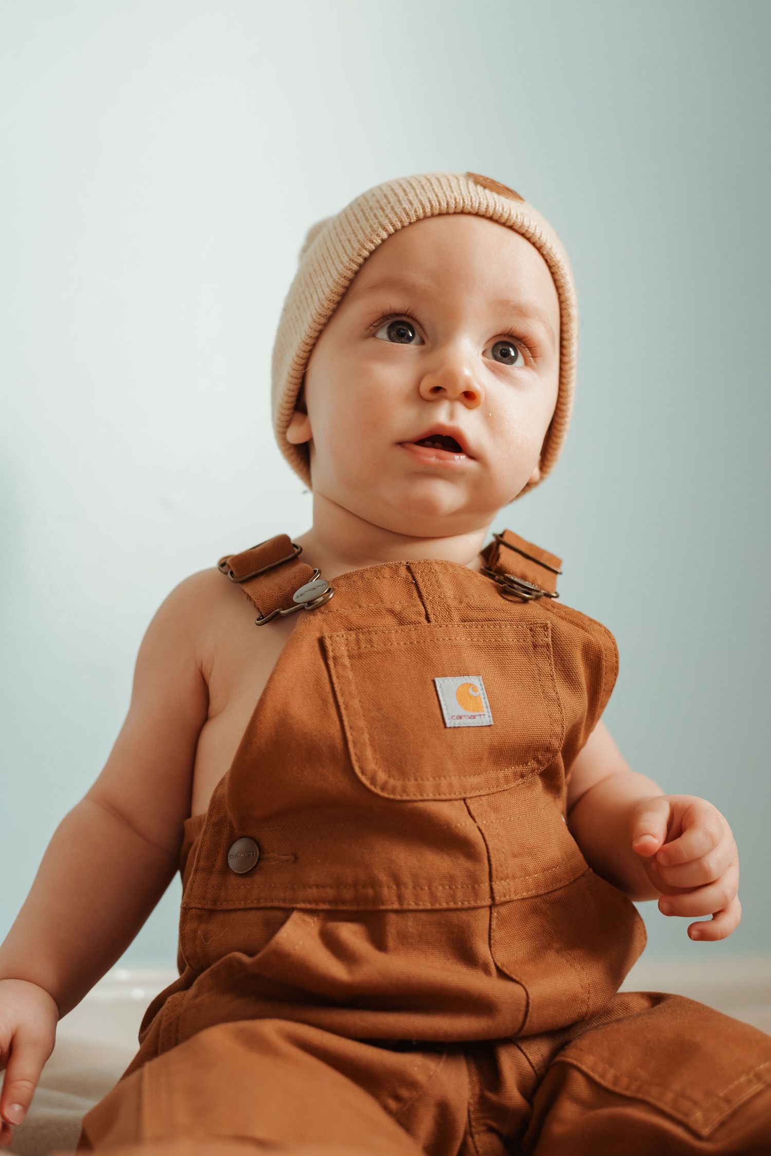 Baby in brown Carhartt overalls and beige knit hat sits looking upward on light background.